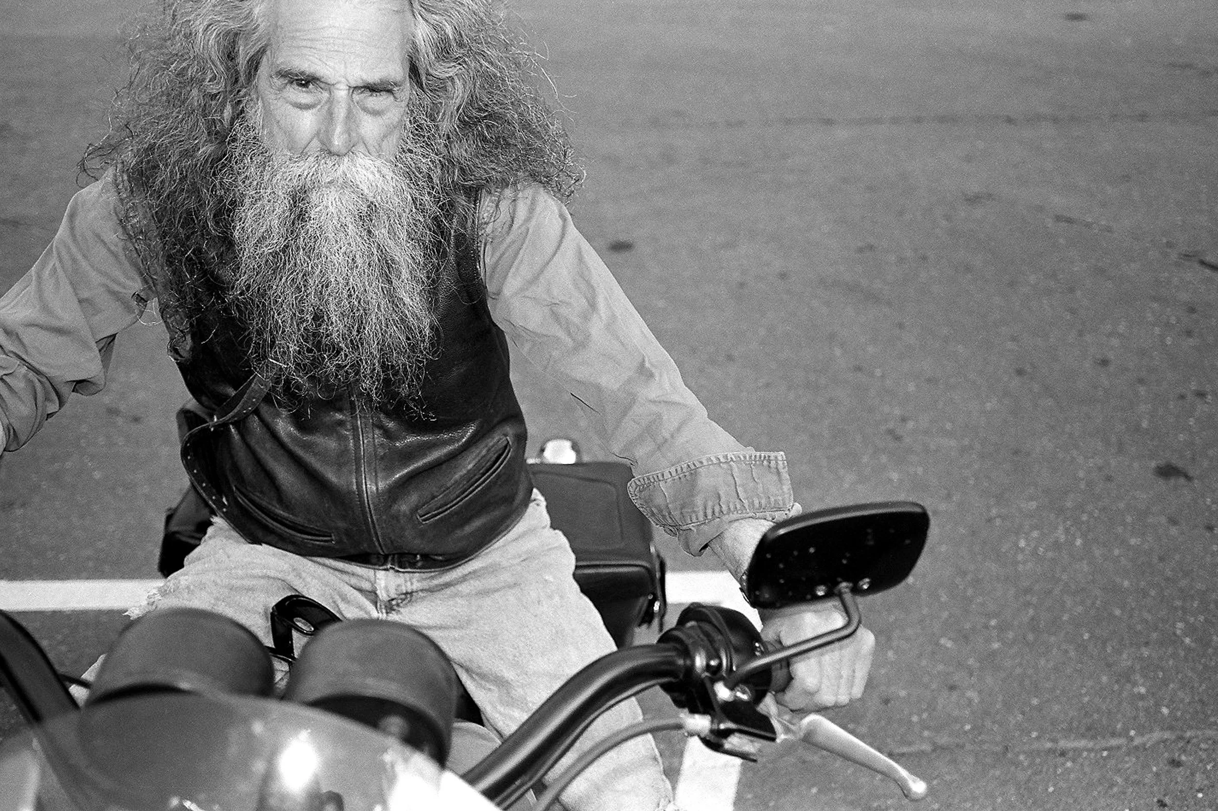 A black-and-white photo of an elderly man with long hair and a beard, riding a motorcycle. The man is wearing a leather vest and sitting on the motorcycle on a street, looking directly at the camera.