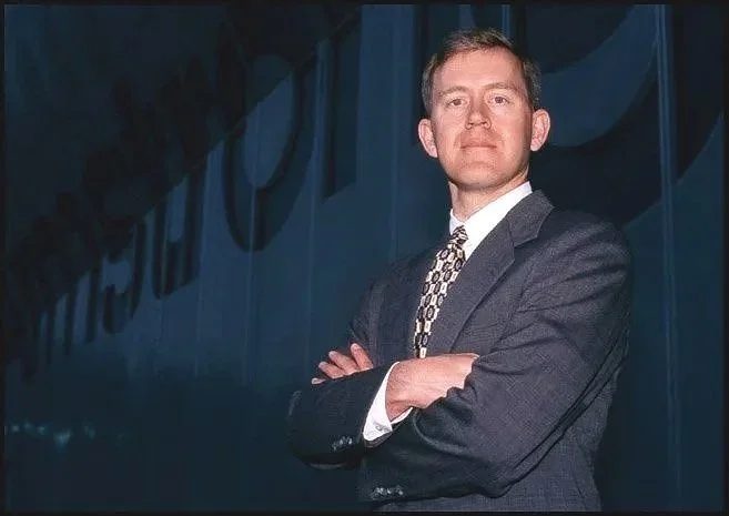 A professional business man in a dark suit with a patterned tie, standing intently with arms crossed in front of a dark wall with company name.