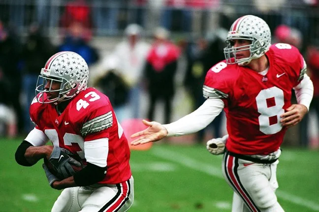 Two American football players from Ohio State, wearing red jerseys with white numbers and helmets, celebrating on the field during a game.