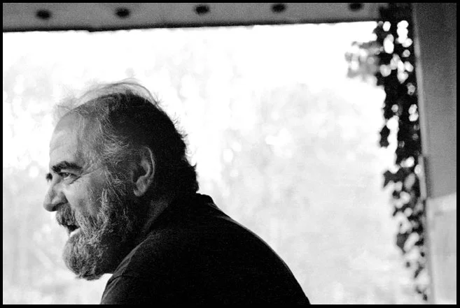 A black-and-white photo of a smiling older man with a beard and short hair, viewed in profile, sitting indoors with a window in the background.