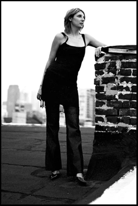 A woman in a black dress and leggings standing on a rooftop with a city skyline in the background, leaning on a chimney.