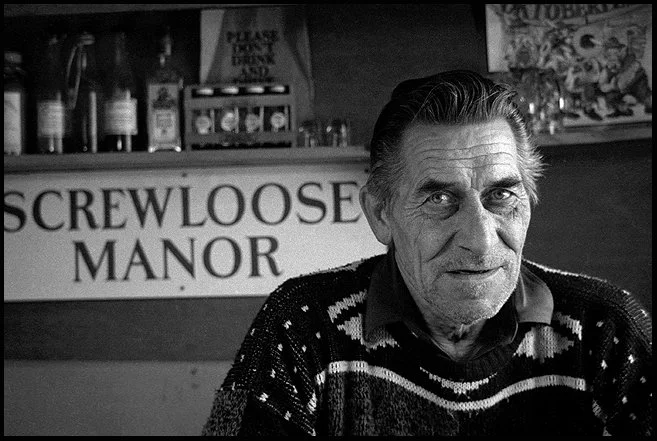 A man sitting in front of a sign that reads 'Screwloose Manor', with shelves holding bottles and boxes in the background.