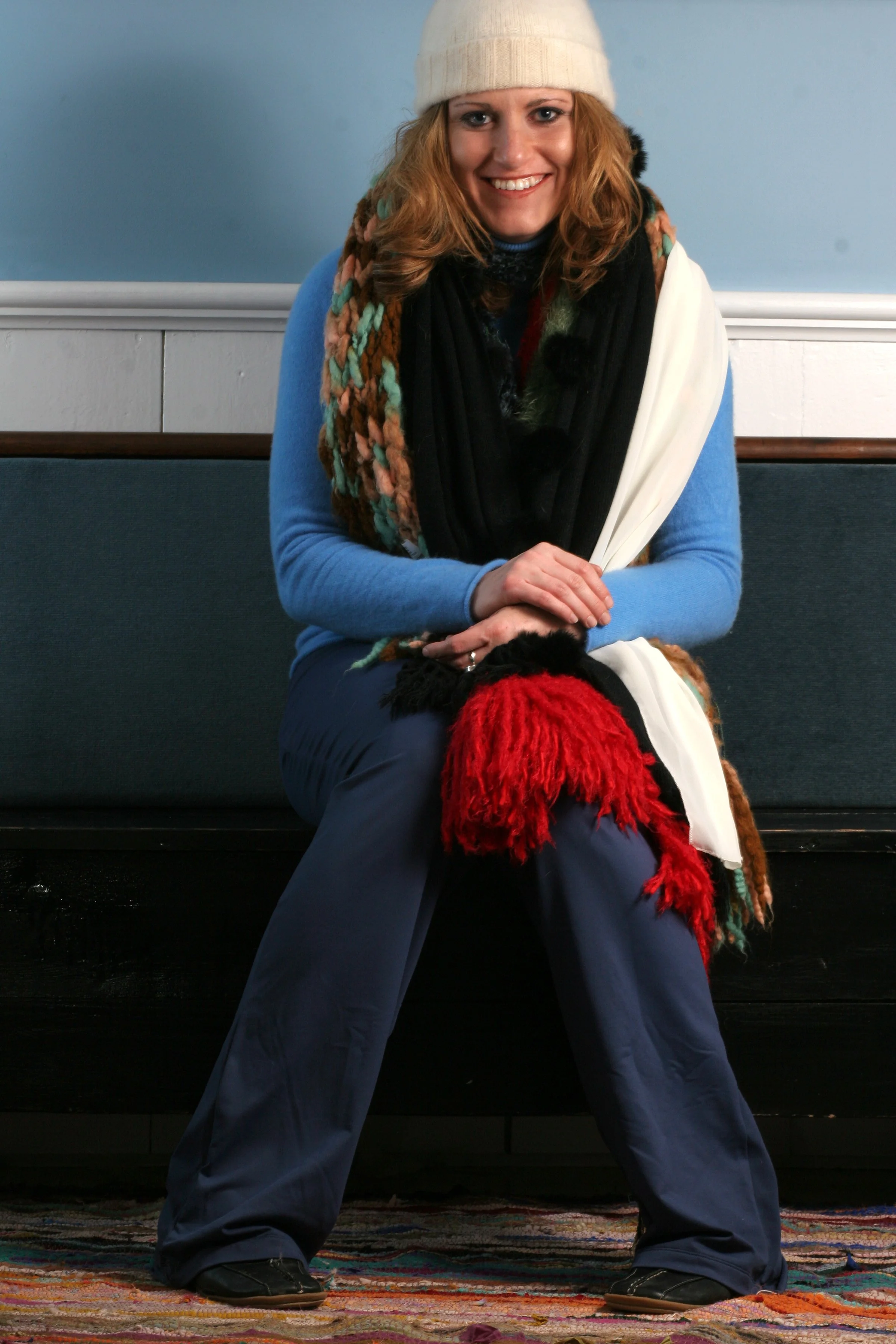 Woman sitting on a bench, smiling, wearing a white beanie, multicolored scarf, blue jacket, and navy pants, with her hands resting on her lap, on a colorful rug indoors.
