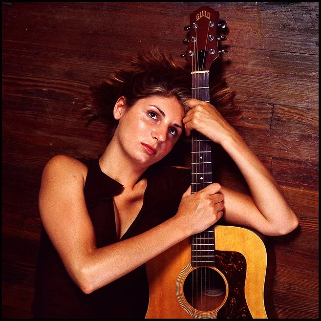 A woman with short, wavy hair lying on a wooden floor, holding an acoustic guitar close to her face.