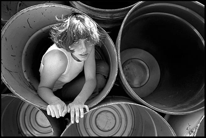 A woman with short, wavy hair sitting inside a large metal pipe, looking up at the camera.