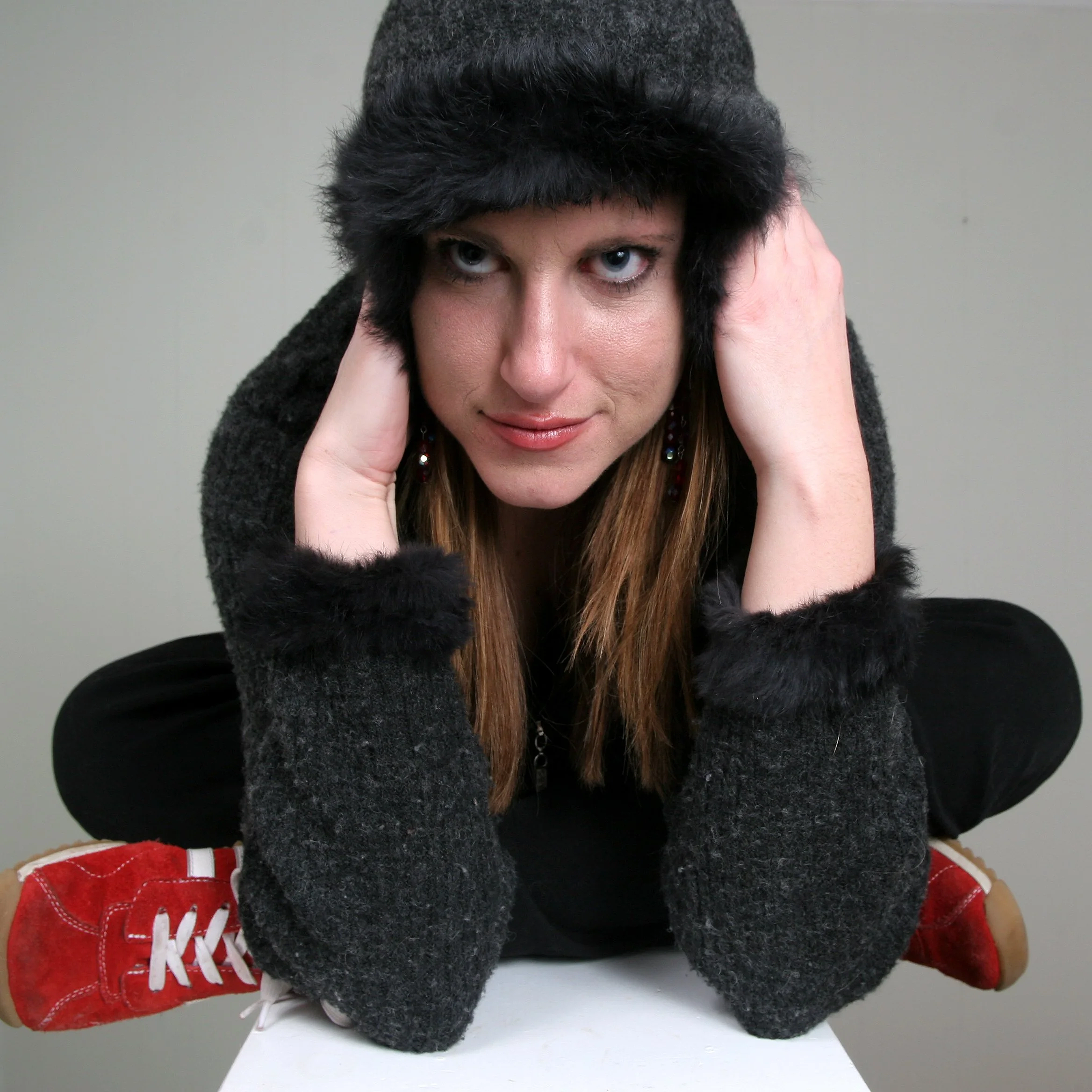 Woman with blue eyes wearing a black fuzzy sweater with hood, holding their head with both hands while sitting cross-leg. They are wearing a dark sweater and red sneakers with white laces. The background is plain and light-colored.
