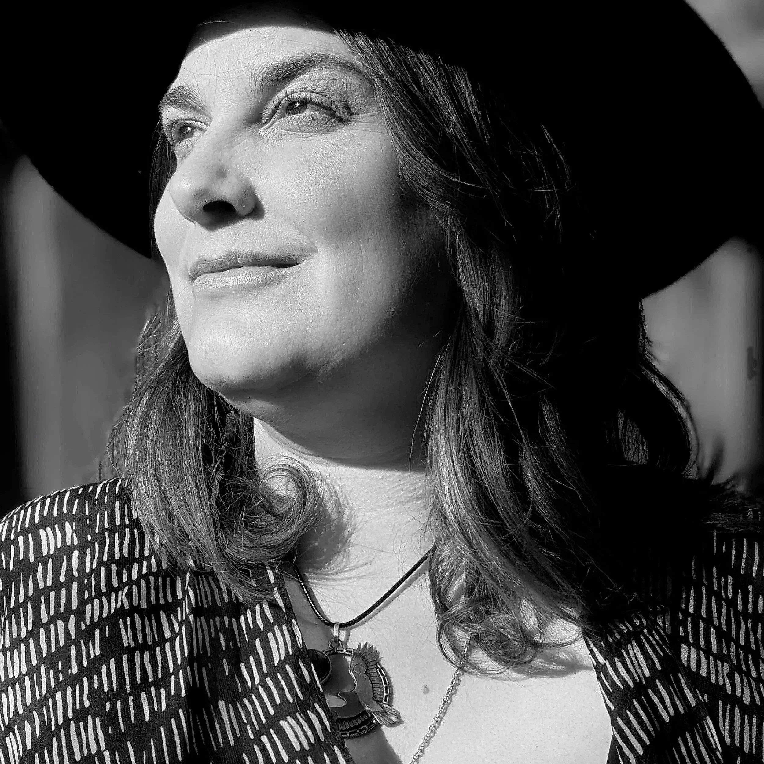 black and white solo portrait of  professional female gazing out toward the distance to the left lit by sunlight wearing large black brim hat