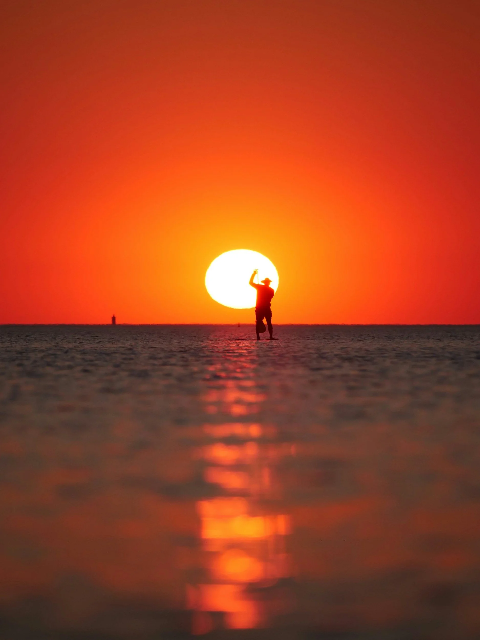 Person standing in shallow water at sunset, with the sun low on the horizon and the sky glowing orange and red
