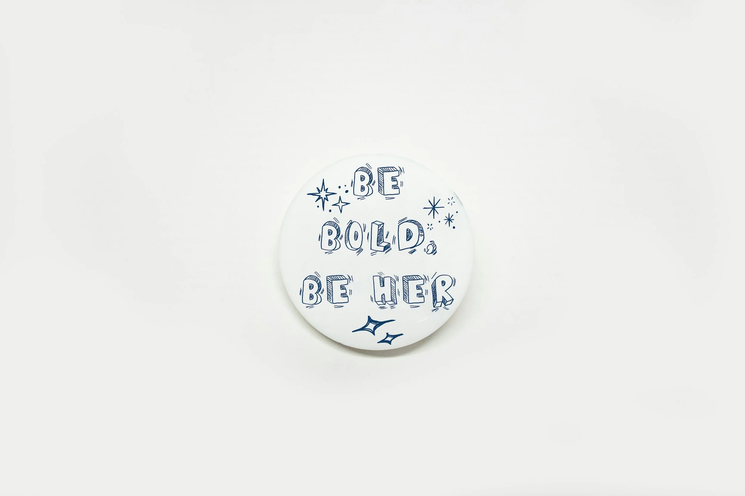 White circular badge with the phrase 'Be Bold, Be Her' written in blue hand-drawn style letters, surrounded by blue stars and sparkles.