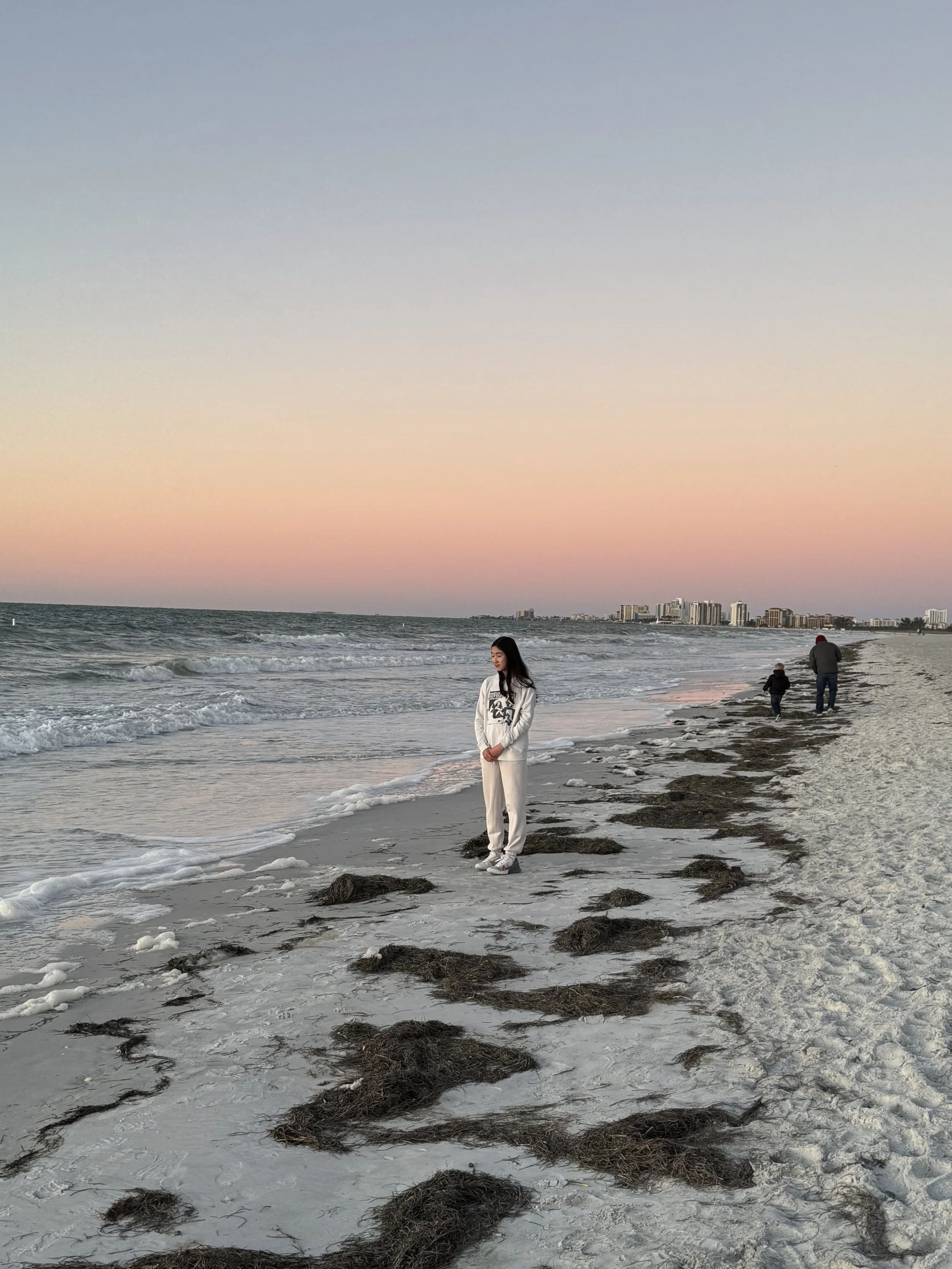 A young woman in light clothing standing on the beach near the shoreline during sunset, with seaweed scattered on the sand and a city skyline in the distance.