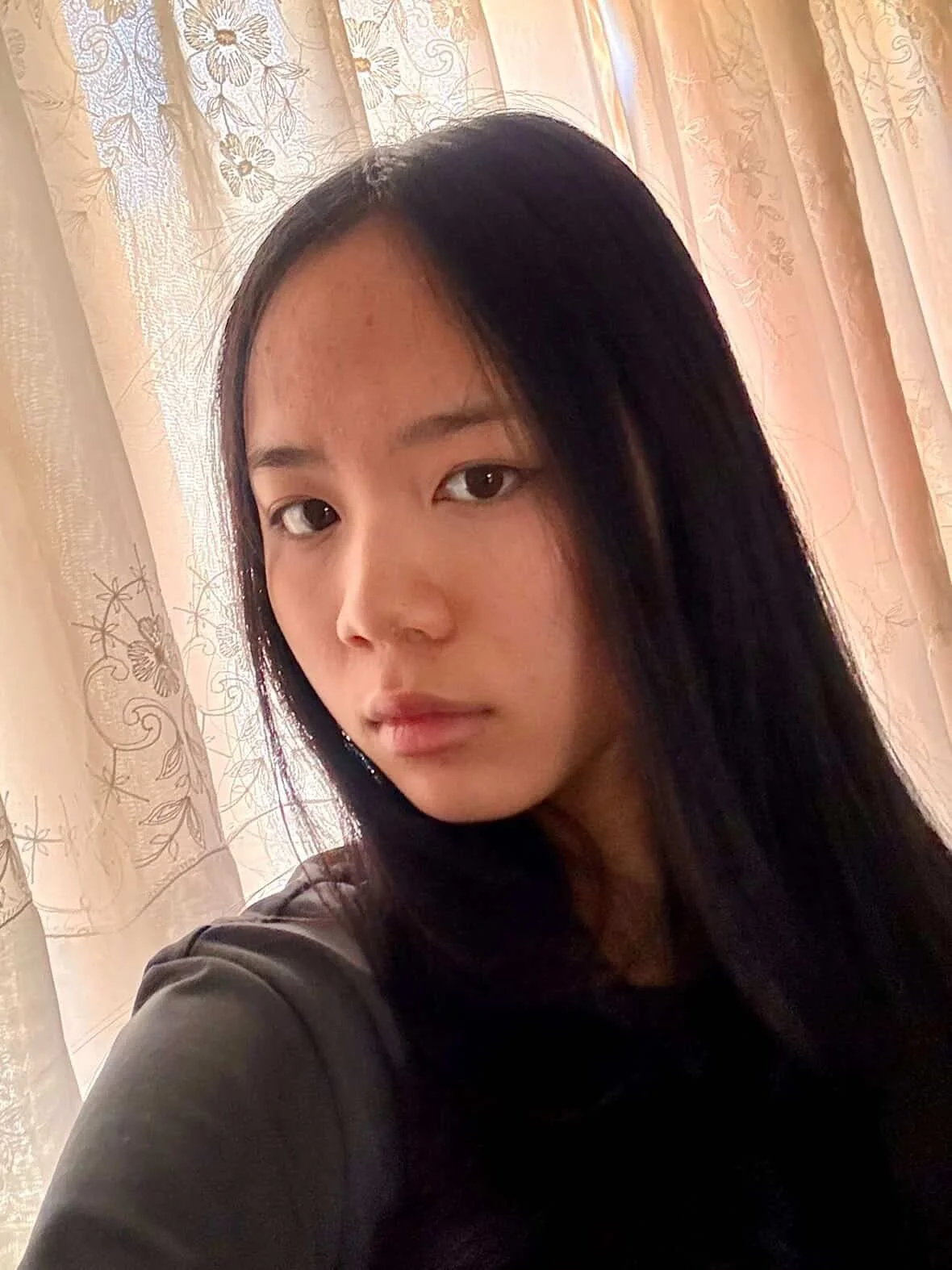 A young woman with long dark hair taking a selfie indoors, with sheer curtains featuring floral and butterfly patterns in the background.