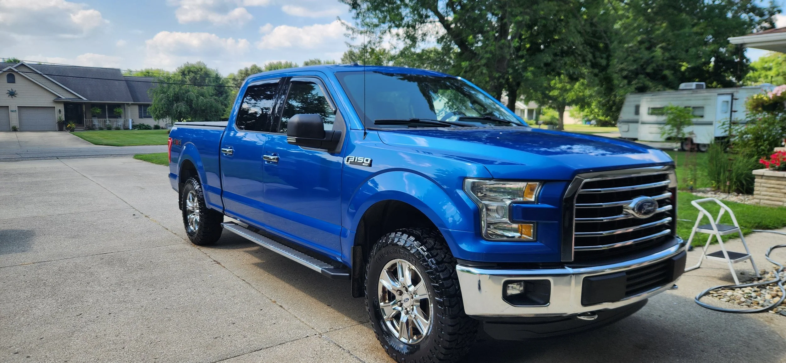 A blue Ford F-150 pickup truck parked on a driveway in a suburban neighborhood with houses, trees, and a recreational vehicle in the background on a partly cloudy day.