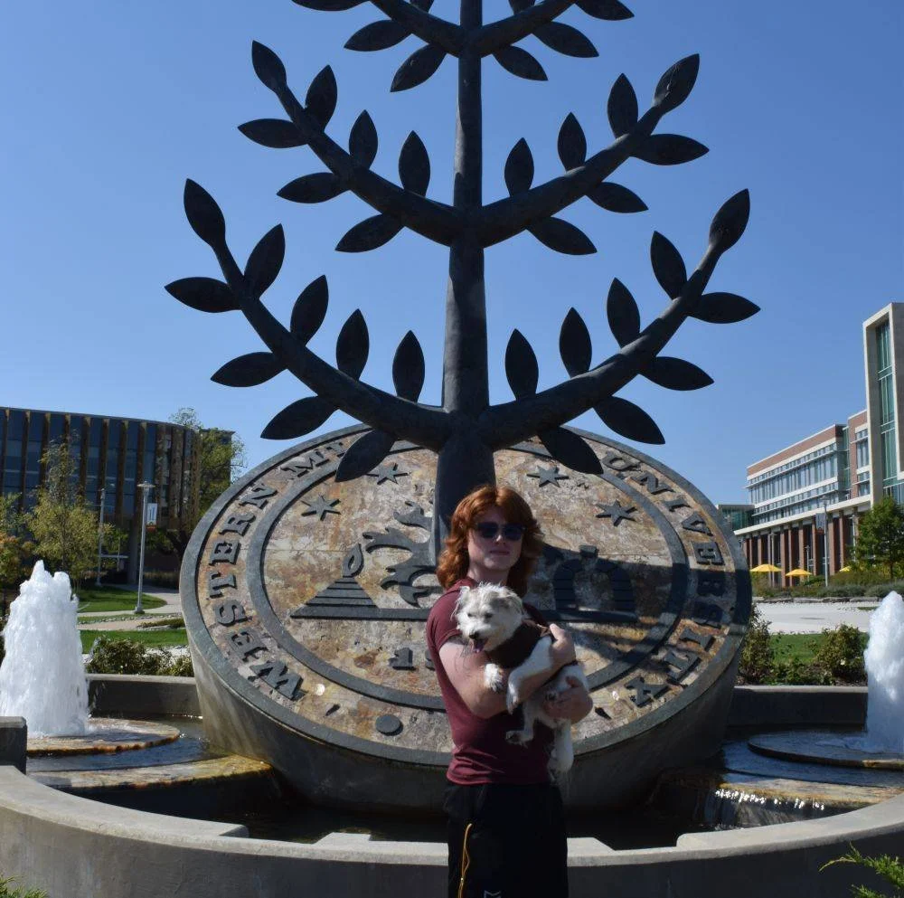 Woman with sunglasses holding a dog in front of a large sundial monument with an artistic tree sculpture on top, in an urban park setting with fountains and modern buildings.