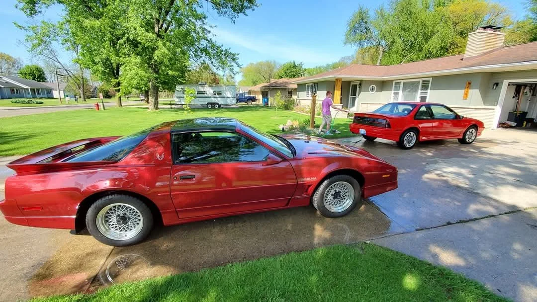 Two red cars parked in a suburban driveway with a woman watering plants nearby. Green lawns, trees, and a camper trailer are visible in the background.