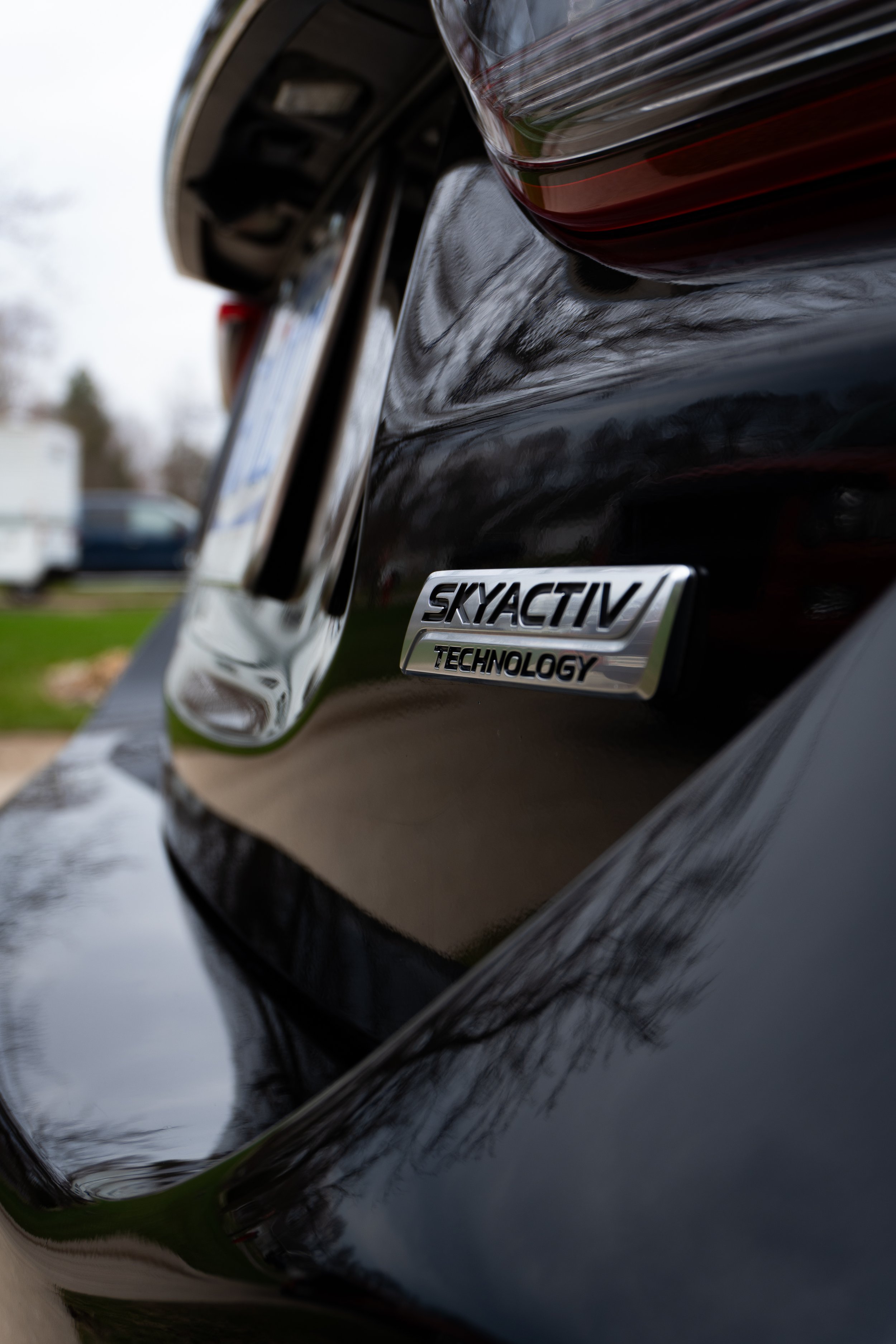 Close-up of the rear of a black vehicle showing a metal badge that reads 'SKYACTIV TECHNOLOGY' and part of the tail light.