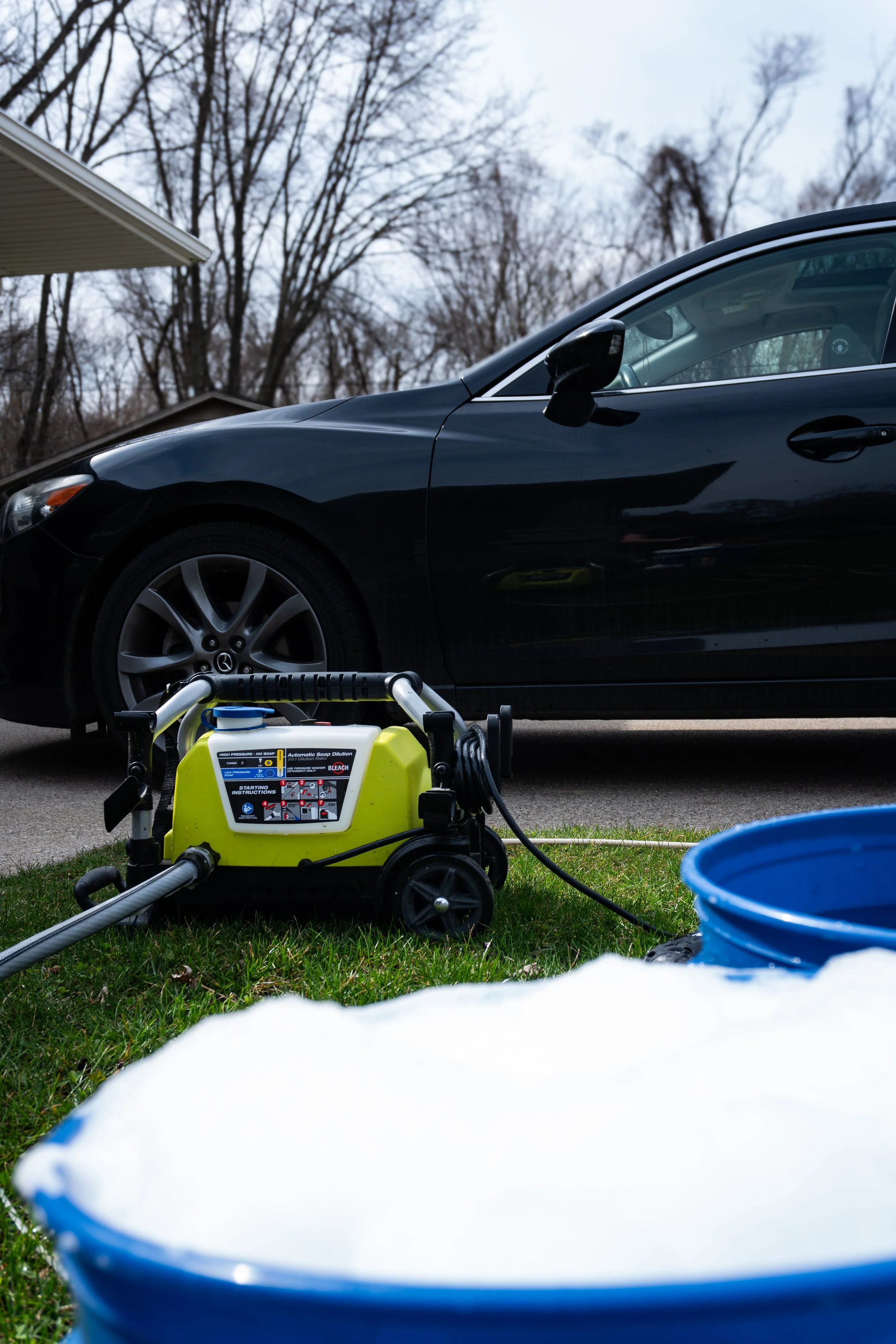 A black car parked outside on a driveway, with a foam washer and blue bucket nearby, suggesting car washing in progress.