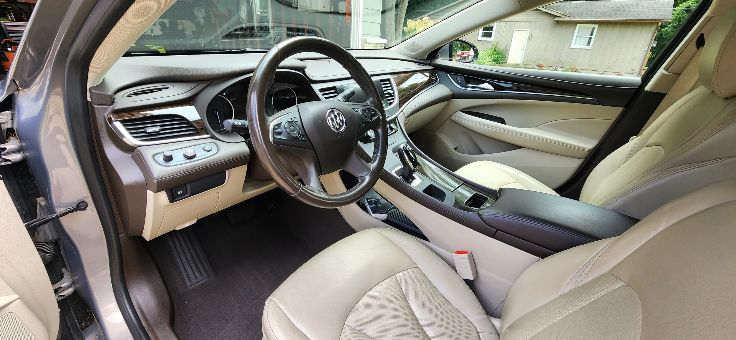 The interior of a luxury car showing the front seats, steering wheel, dashboard, center console, and gear shift, with a view outside the windshield of a house and greenery.