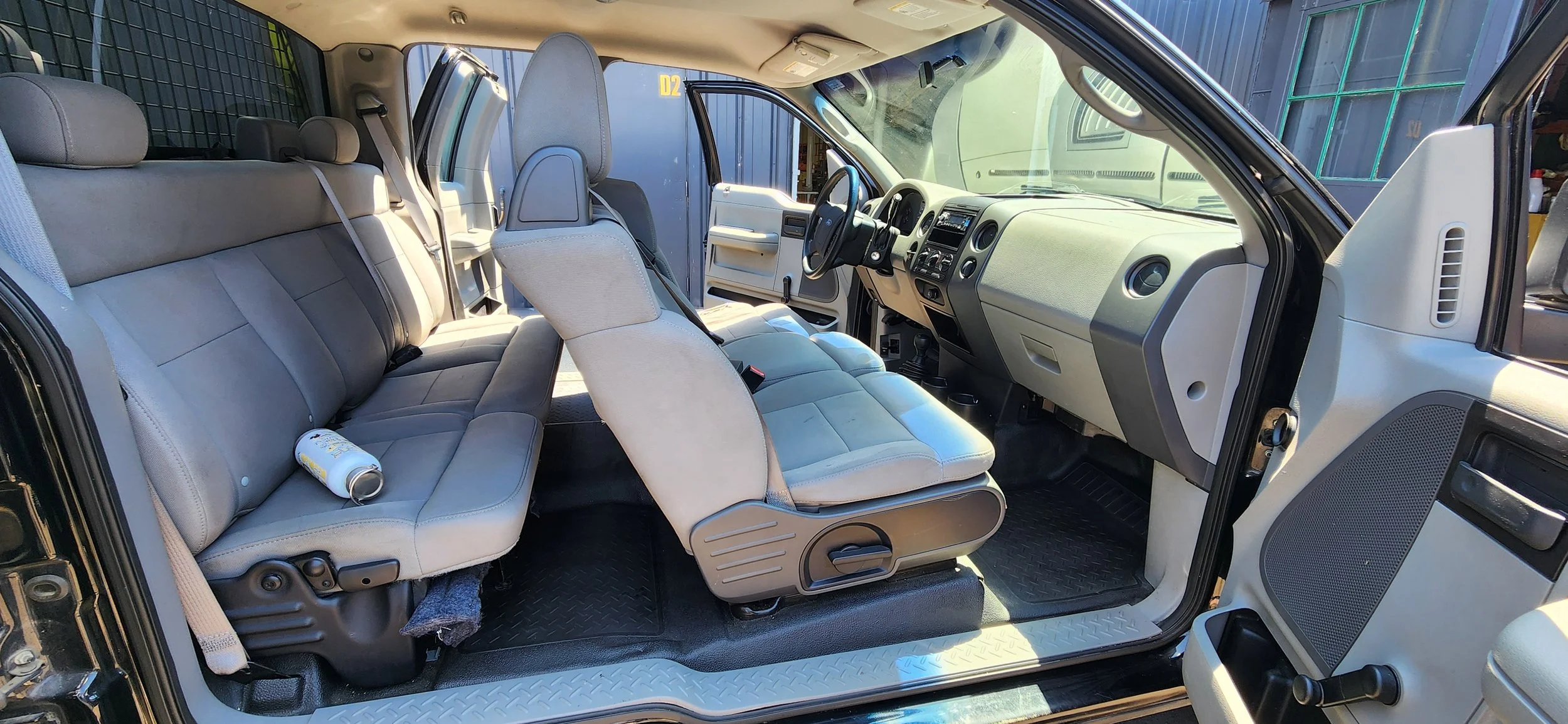 Interior of a vehicle with gray seats, the front seats and a soda can on the rear seat, dashboard, steering wheel, and open driver's side door.
