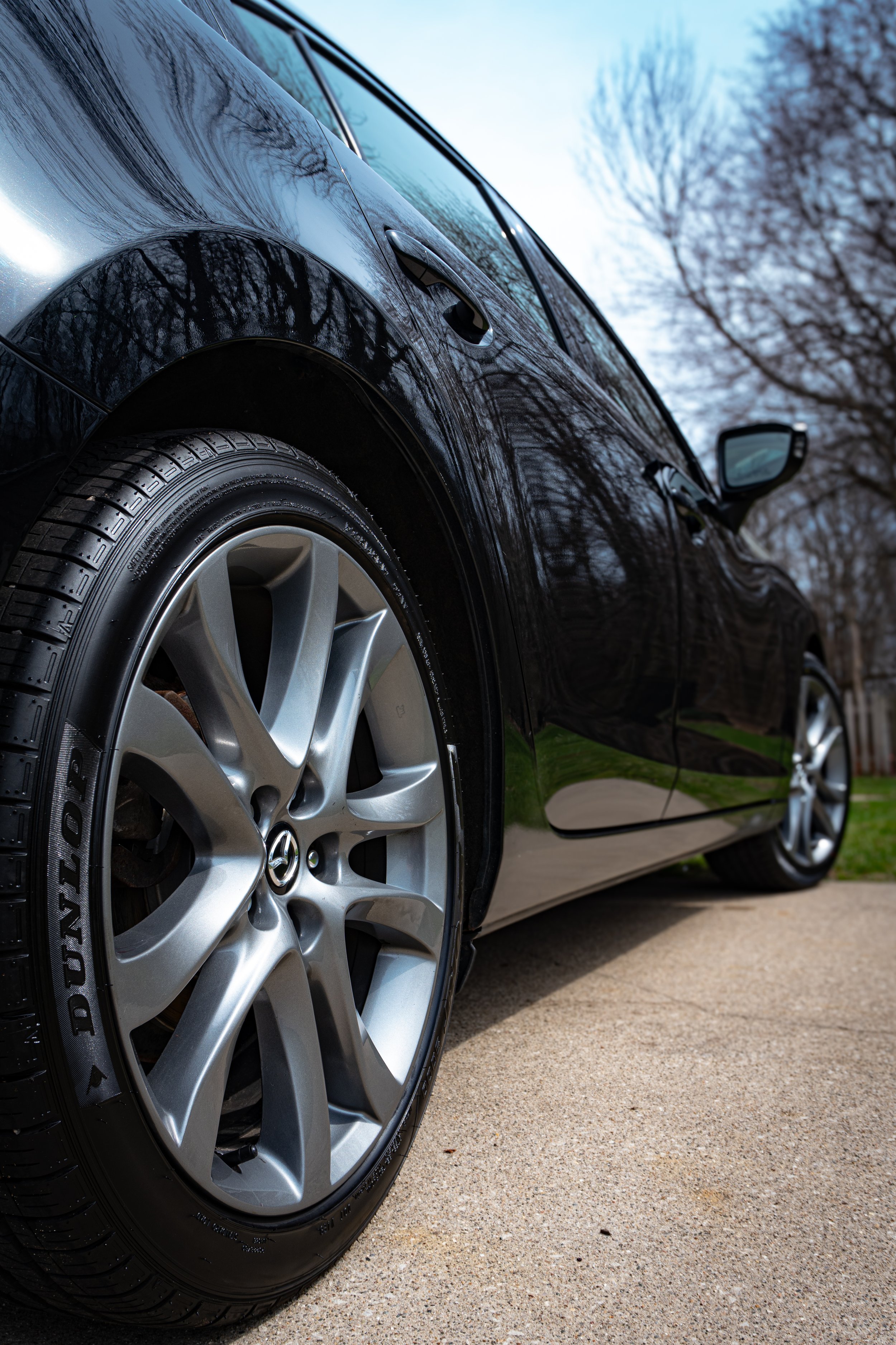 Close-up of a black car's front wheel and part of the side, parked on a driveway with a background of trees and a blue sky.