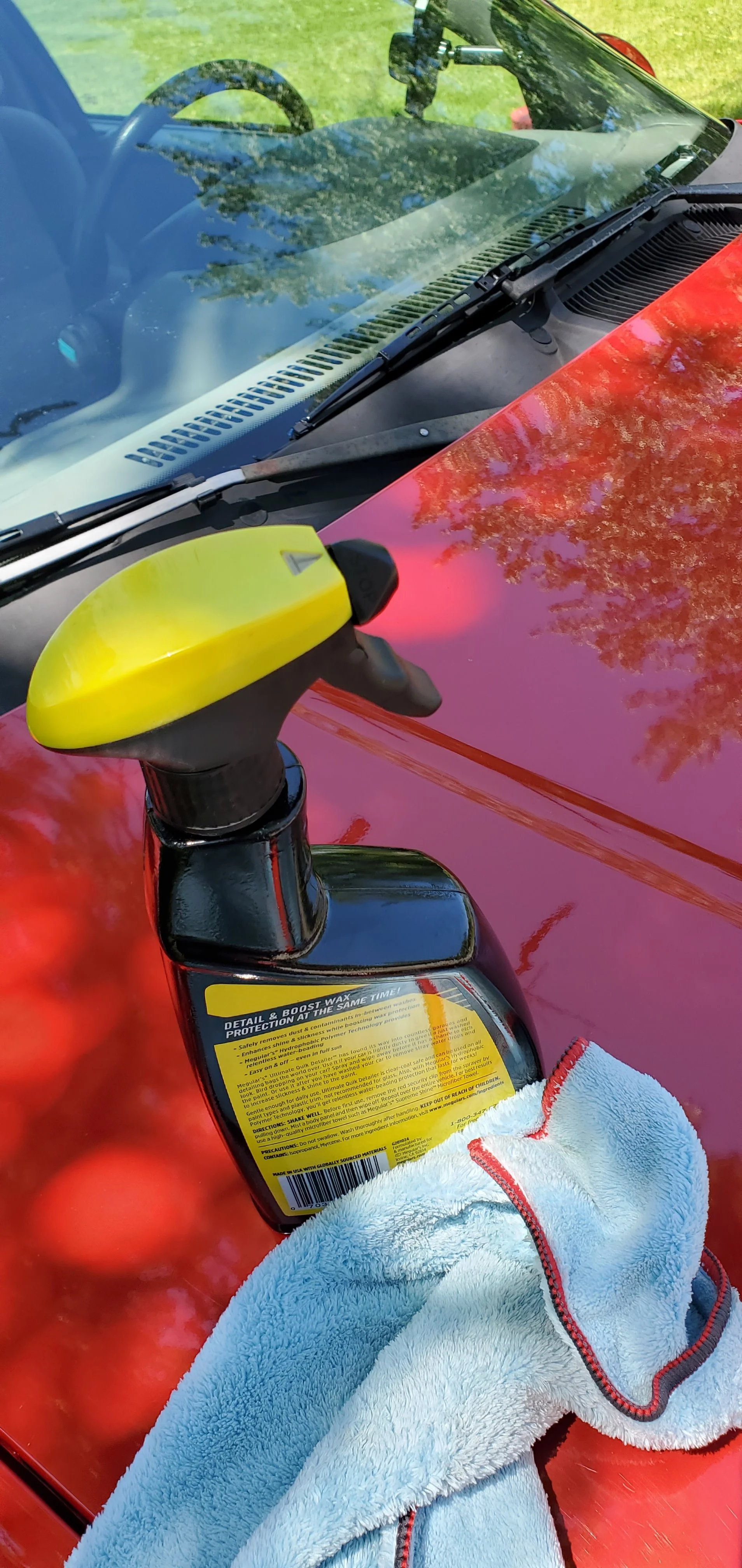 A yellow and black spray bottle labeled 'Detail & Boost Wax' resting on the hood of a red car, with a white towel nearby, and the car's windshield reflecting trees and the sky.