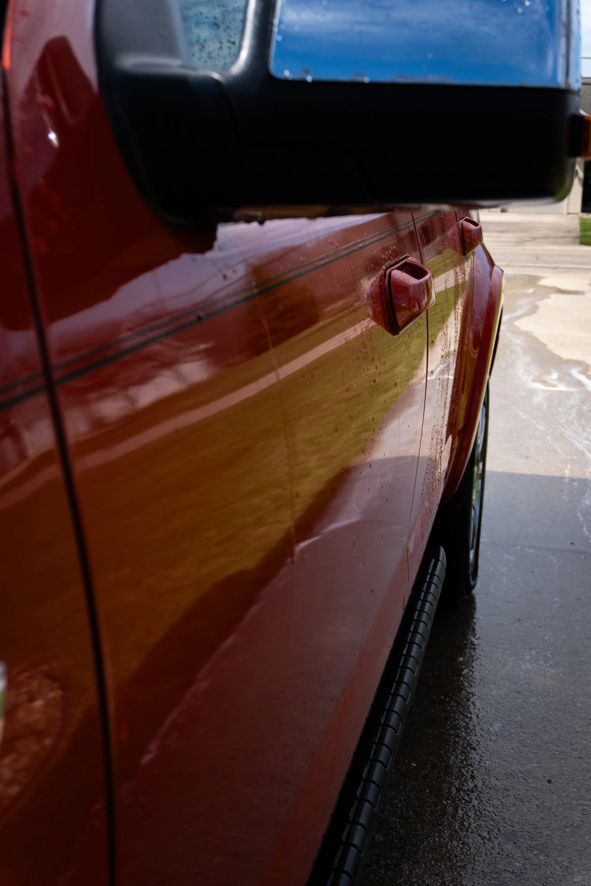 Close-up of a red vehicle side, showing a side mirror and door handle, with reflections on the wet surface.