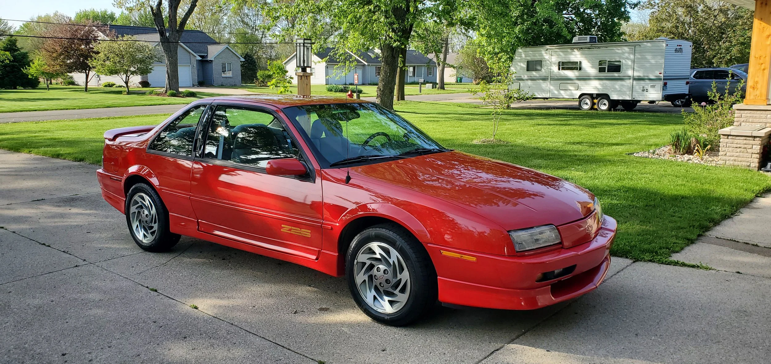 A red sports coupe parked on a driveway in front of a suburban yard with green grass and trees, with a white travel trailer and parked cars in the background.