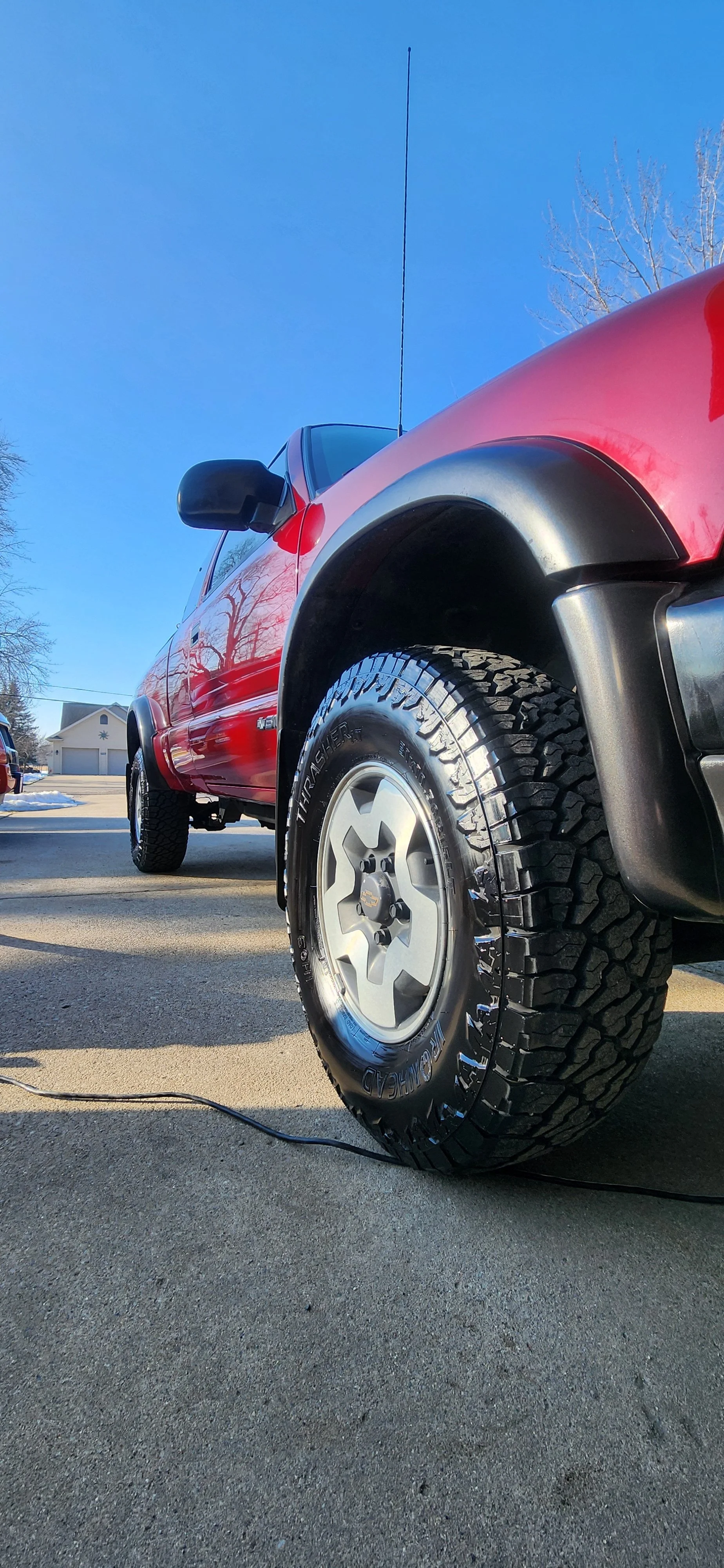 Close-up of a red pickup truck with black side mirrors, parked on a driveway under a clear blue sky. The truck has large off-road tires and a Chevrolet logo on the hubcap. There is a black cable on the driveway and a white house with a garage in the background.
