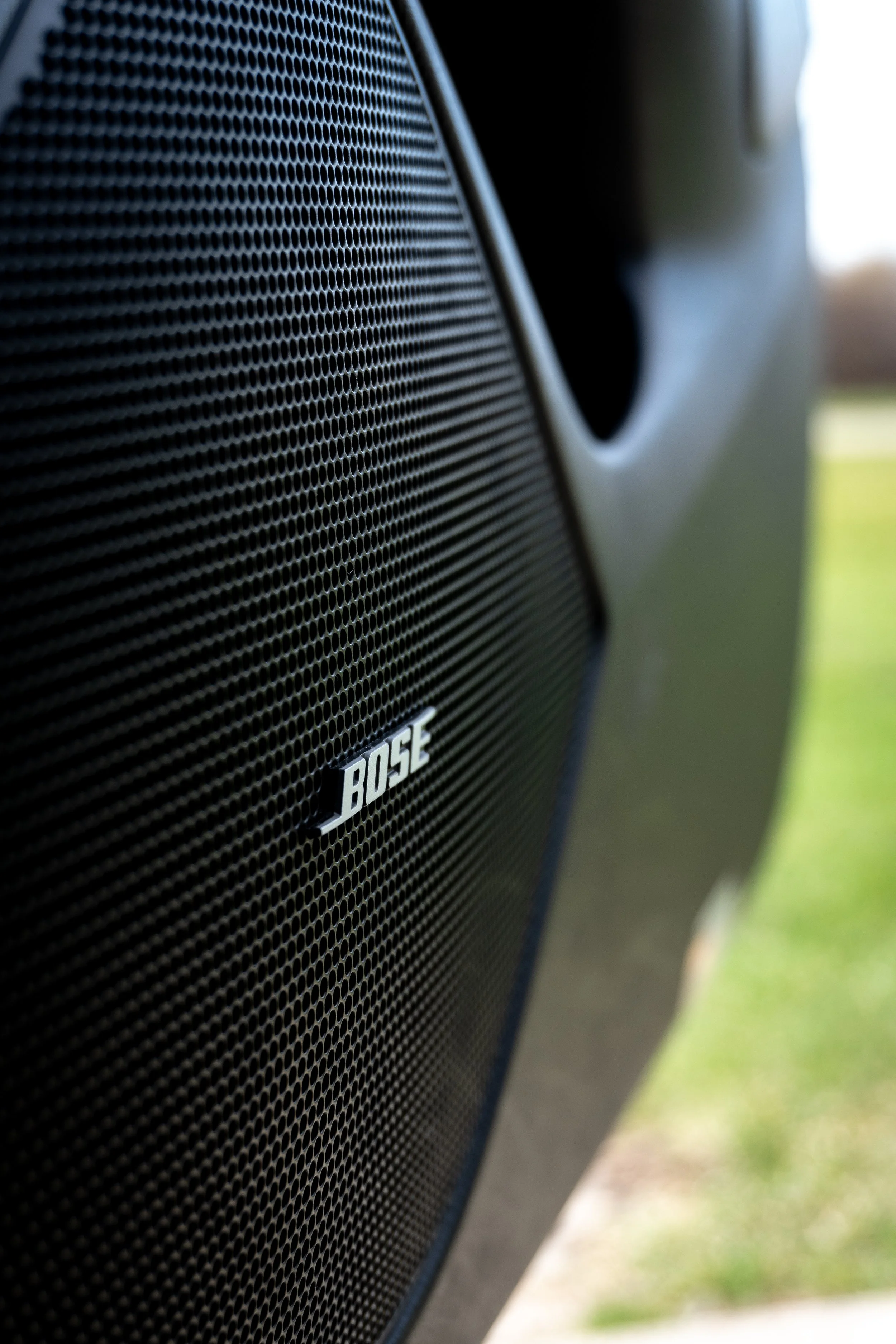 Close-up of a black Bose speaker grille mounted on a vehicle, with grass and sky in the background.
