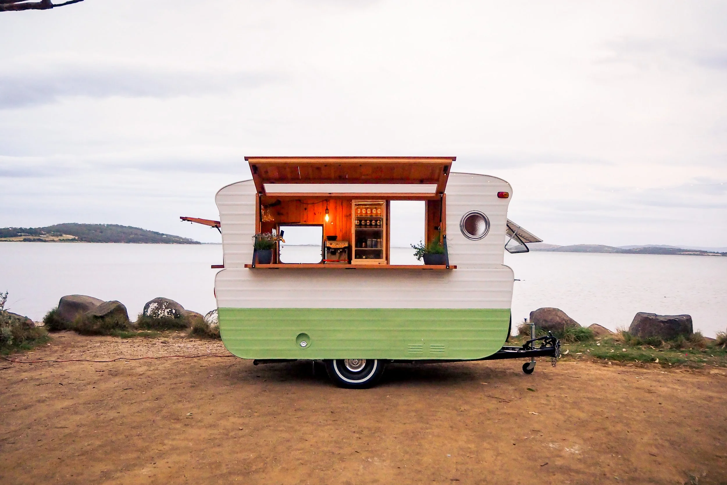A vintage camper trailer with a kitchen setup inside, parked on a dirt area by a body of water with hills in the background.