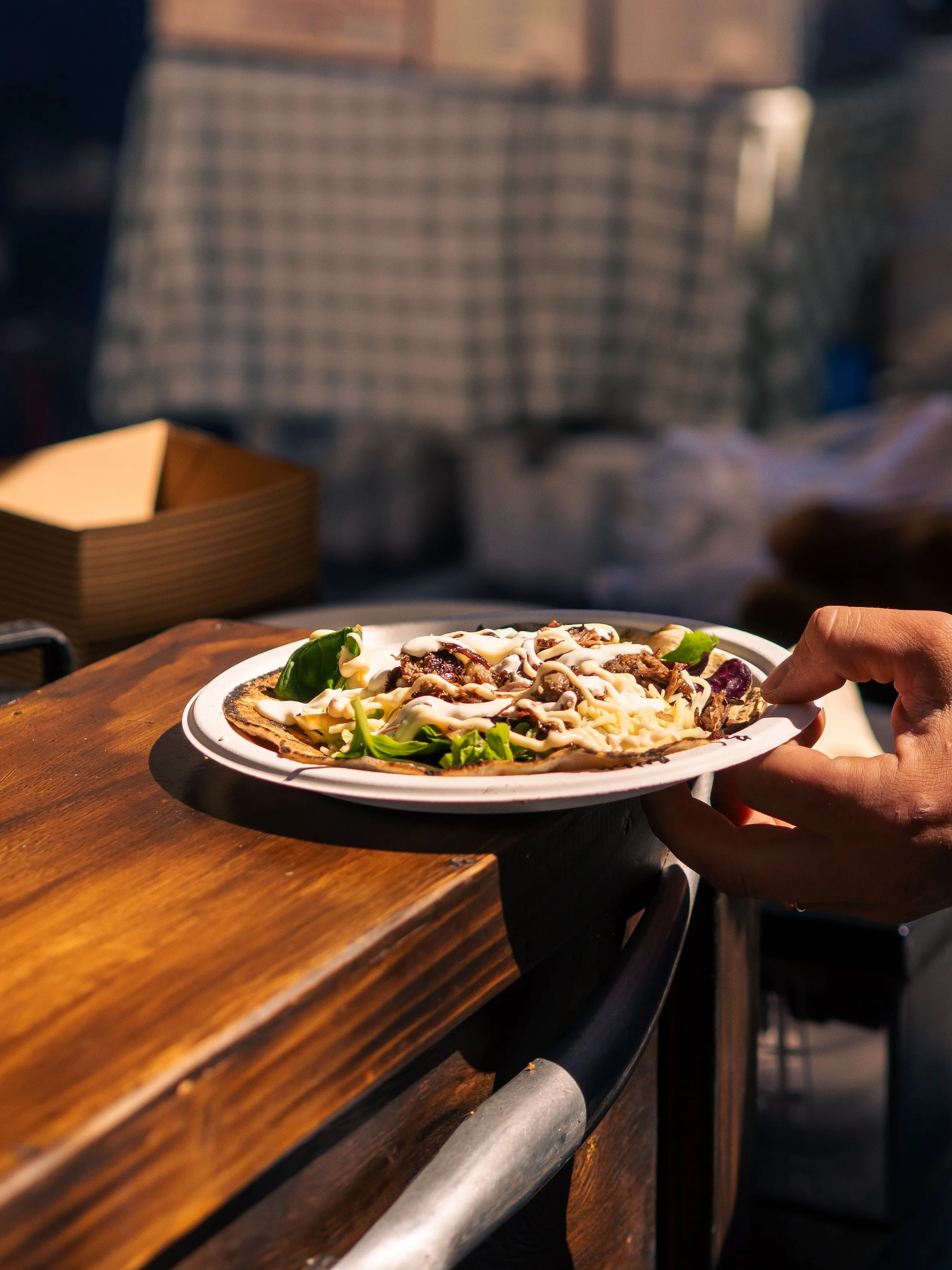 A person holding a plate of food, possibly a pizza, with toppings and drizzled sauce, sitting at a wooden table in a restaurant or outdoor dining area.
