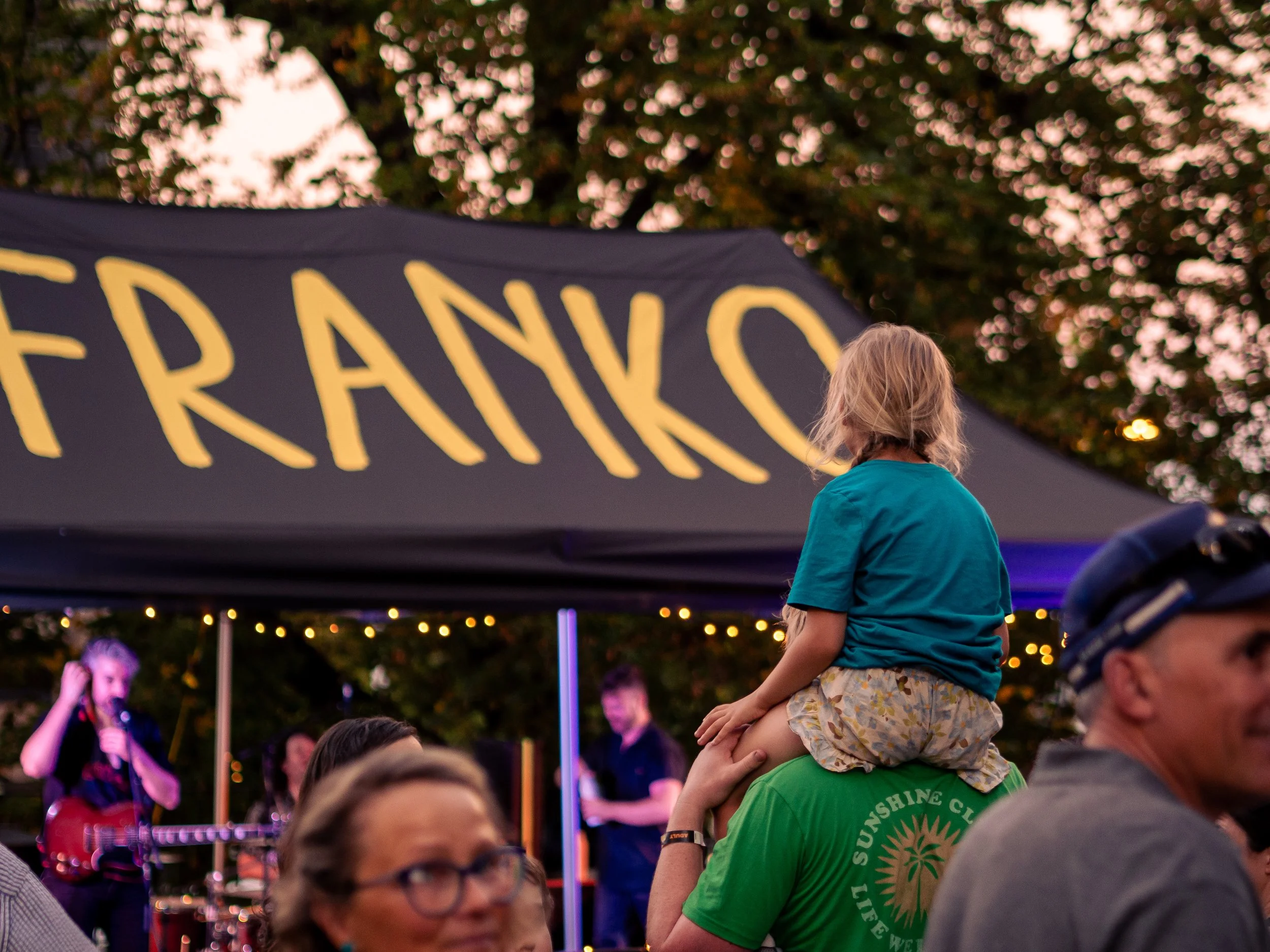 A young girl with blonde hair wearing a teal shirt and floral shorts sitting on a person's shoulders at franko street eats in hobart tasmania, with a crowd of people, a large banner, and trees with pink blossoms in the background during dusk.