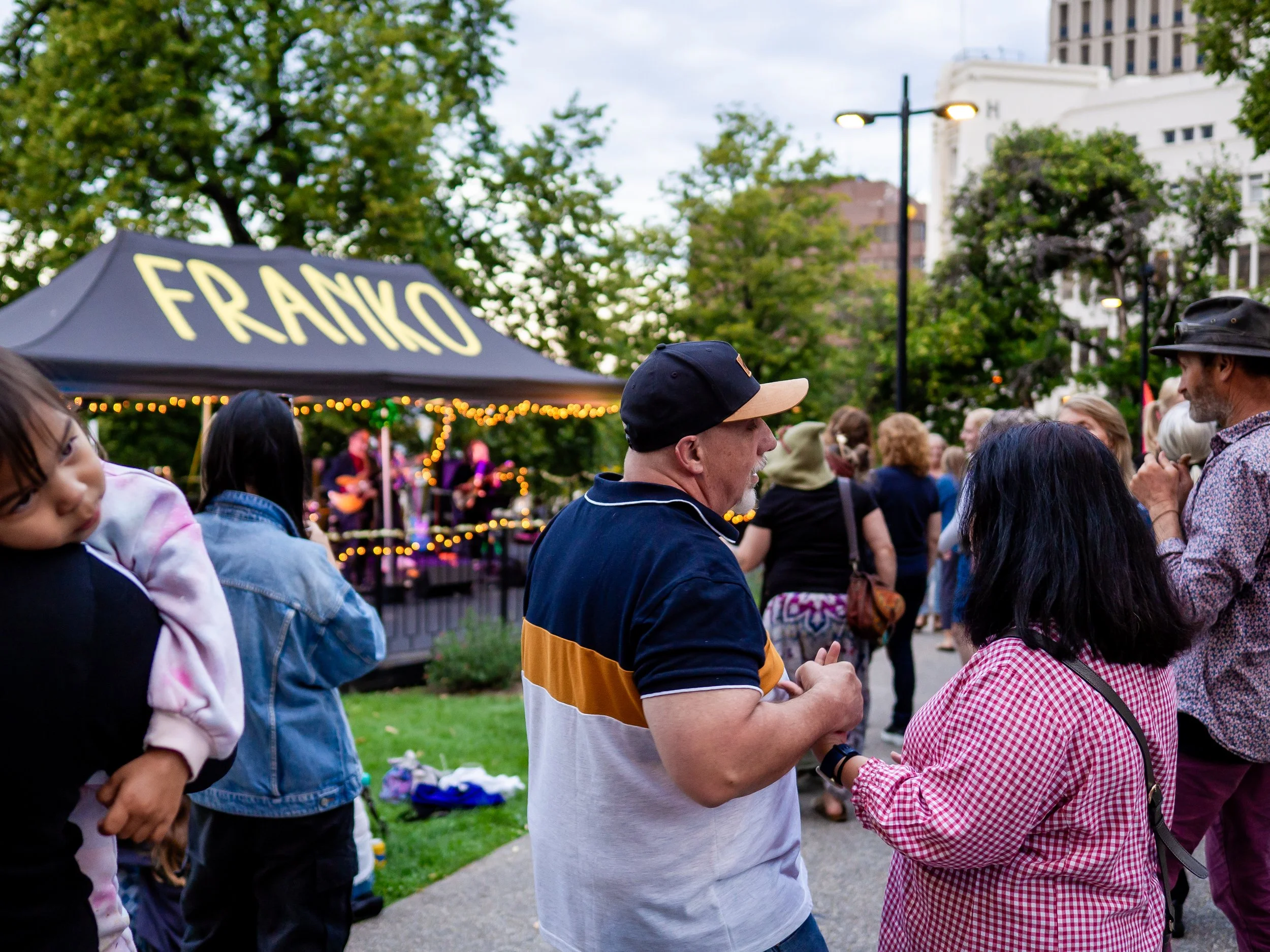 People gathered outdoors during an event in a park with a stand labeled 'FRANKO,' trees, and buildings in the background. Some individuals are engaged in conversation, while a band performs on a stage.
