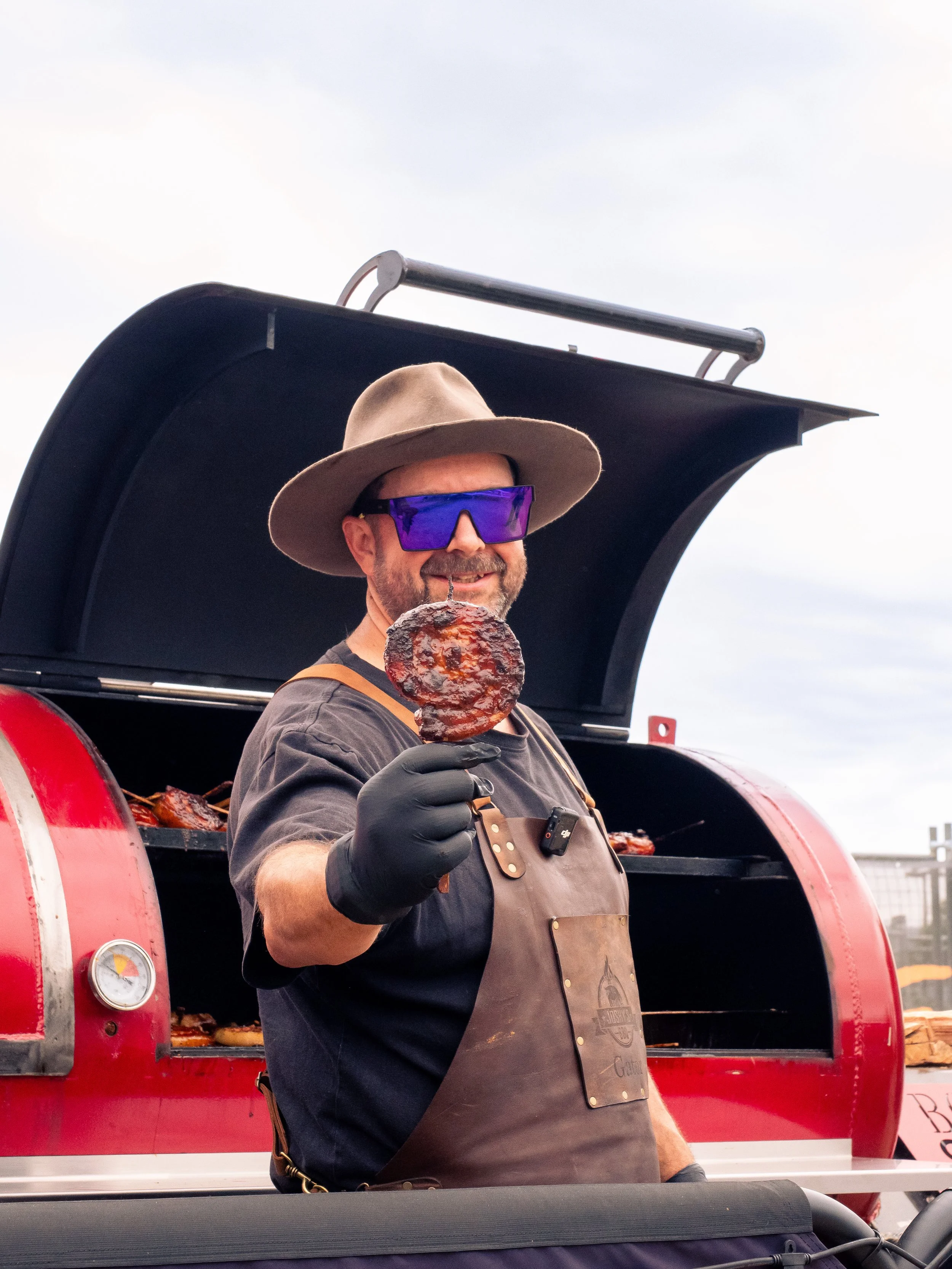 A man wearing a hat and purple sunglasses is holding a piece of grilled meat in front of a red barbecue smoker machine, smiling at the camera.