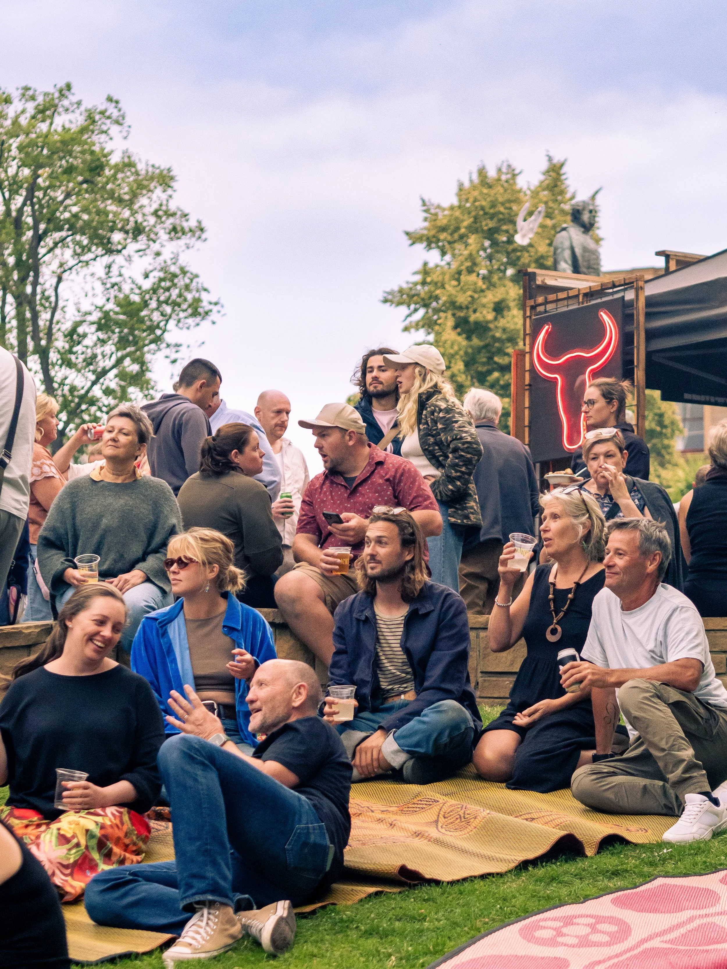 People gathered outdoors on a grassy area during daytime, sitting on mats and socializing. Some are holding drinks, and there is a black sign with a red neon bull head outline visible.