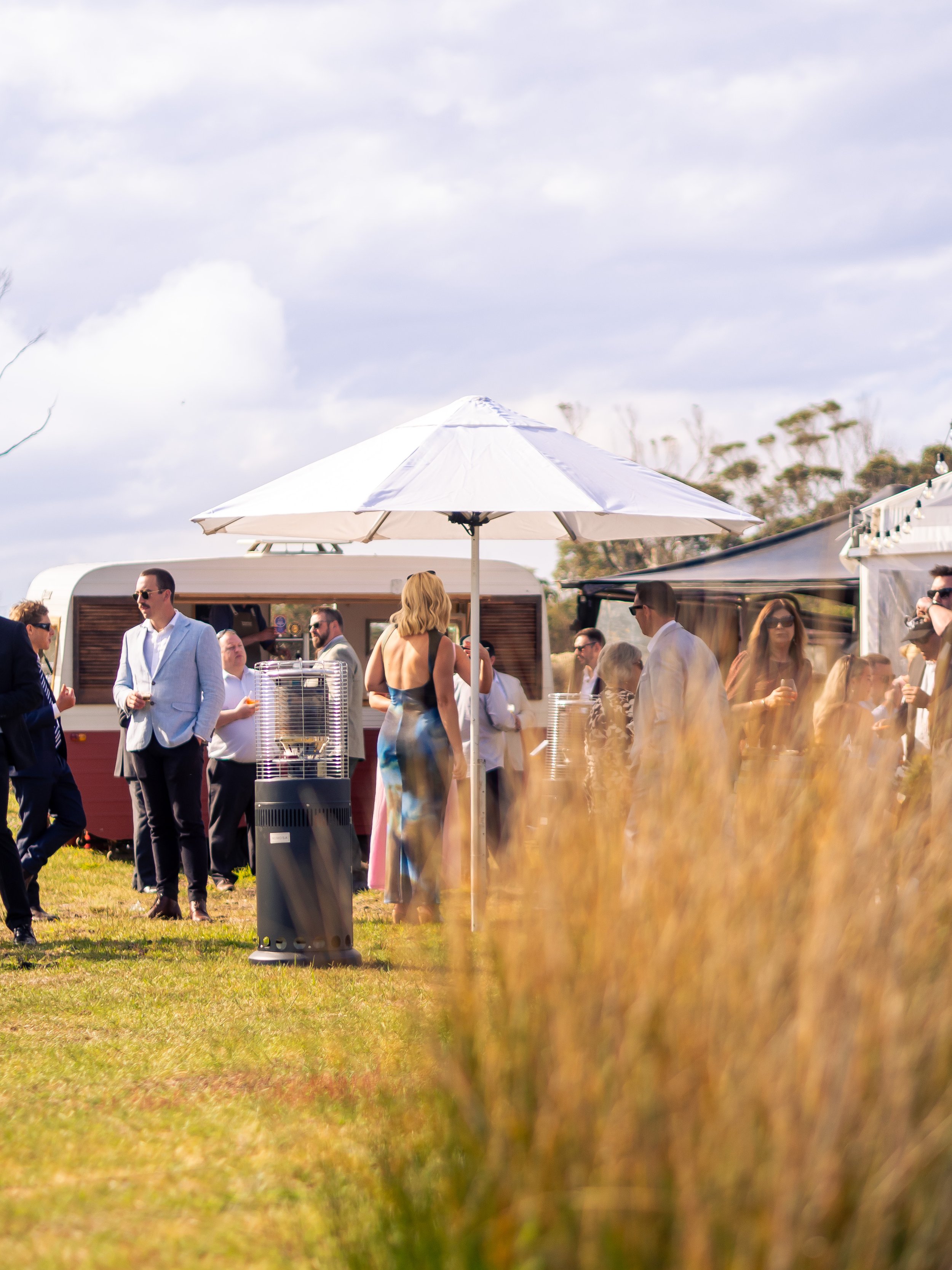 People gathering outdoors at an event with a white parasol, food trucks, and a grassy area, under a cloudy sky.