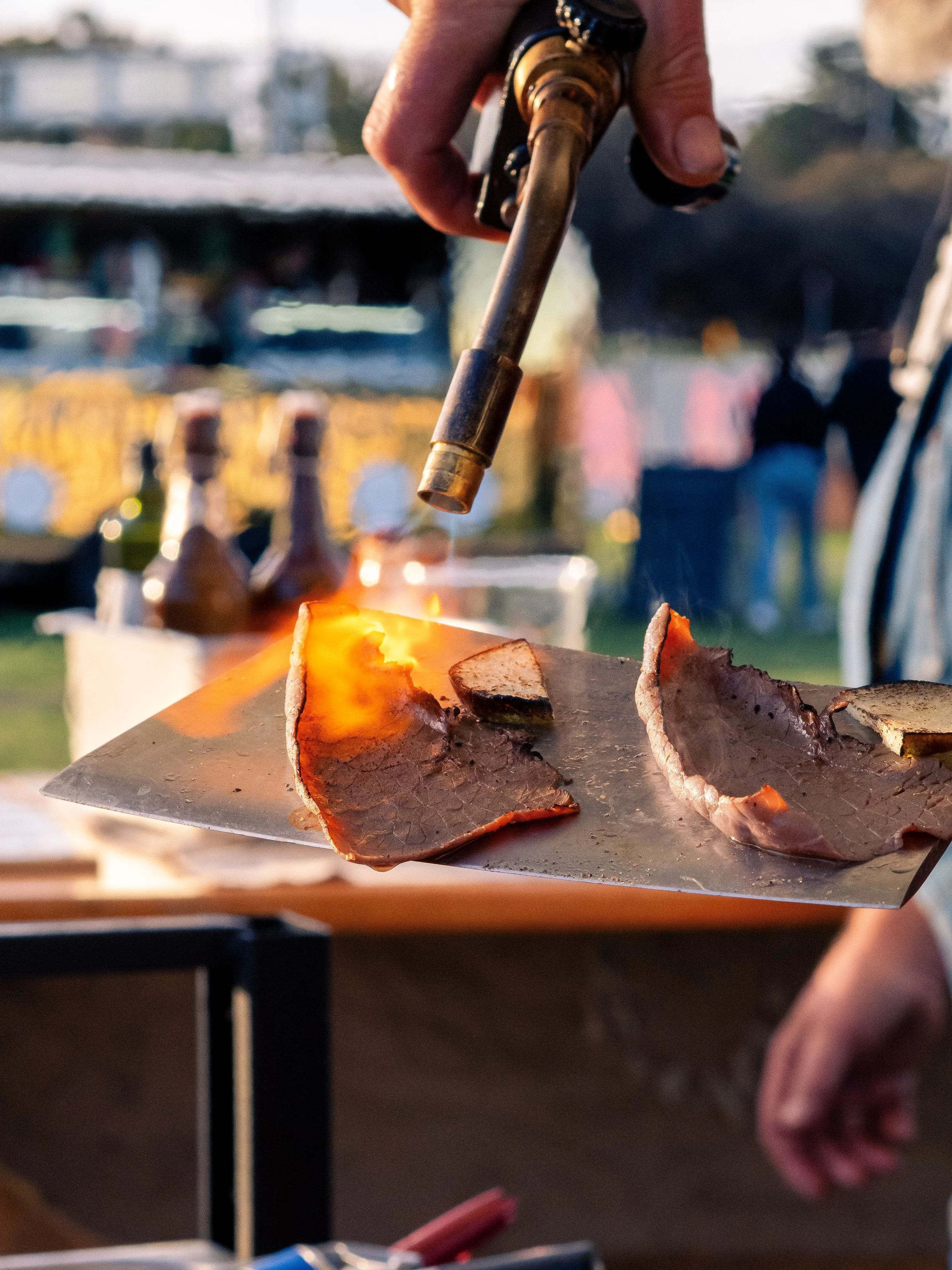 A person is holding a spatula and flipping slices of cooked meat over a grill. The background shows bottles, possibly condiments or drinks, on a table, with blurred outdoor setting.
