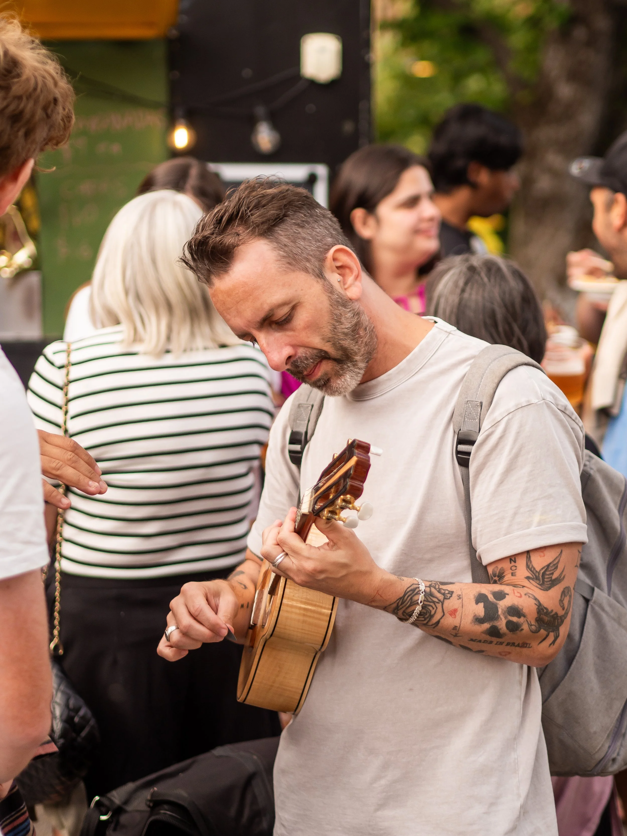 A man with tattoos playing a small guitar in a crowded outdoor setting.
