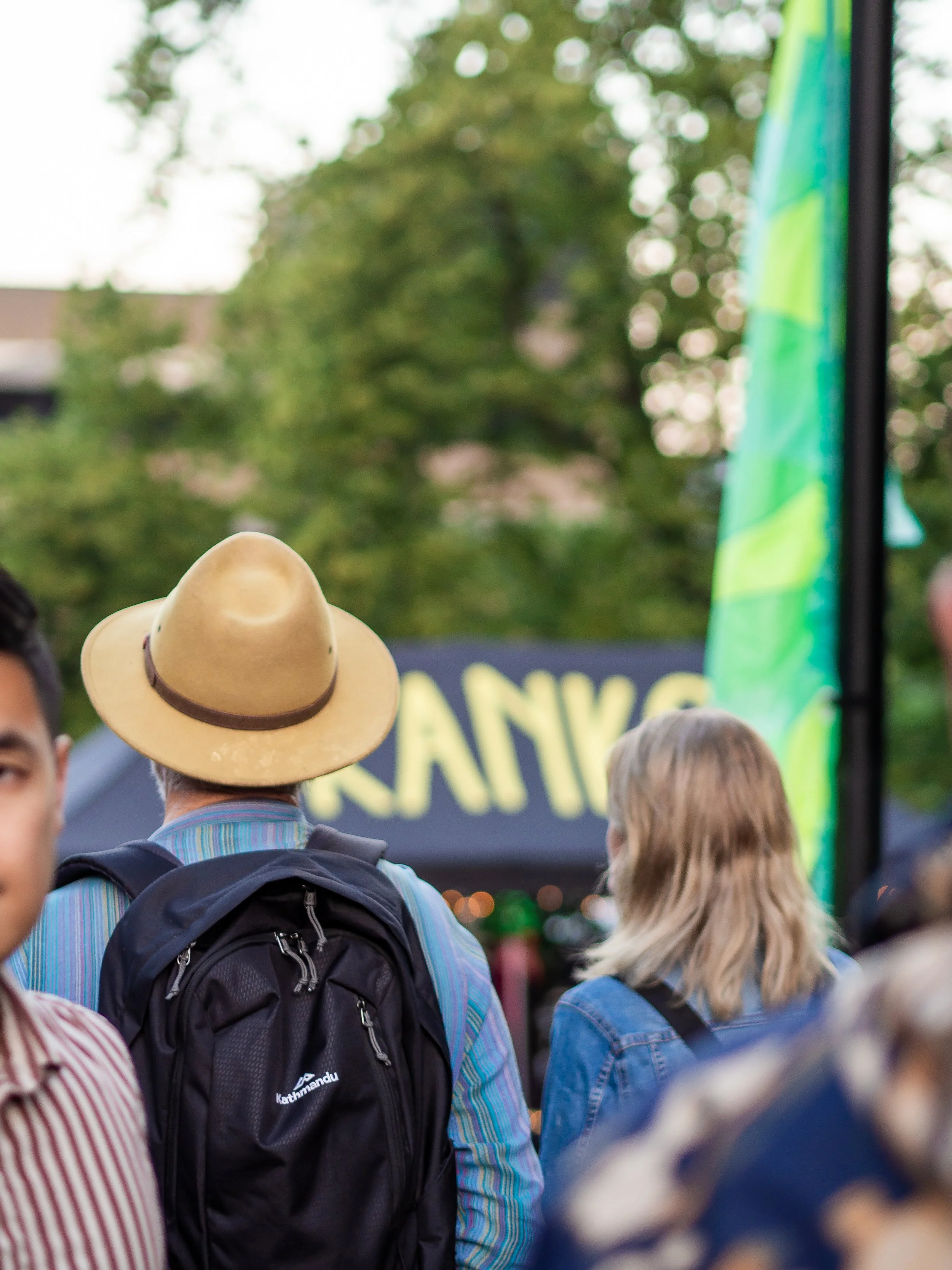 Crowd of people gathered outdoors, with a person wearing a straw hat and another person with blonde hair visible from behind. There are trees and blurred signage in the background.