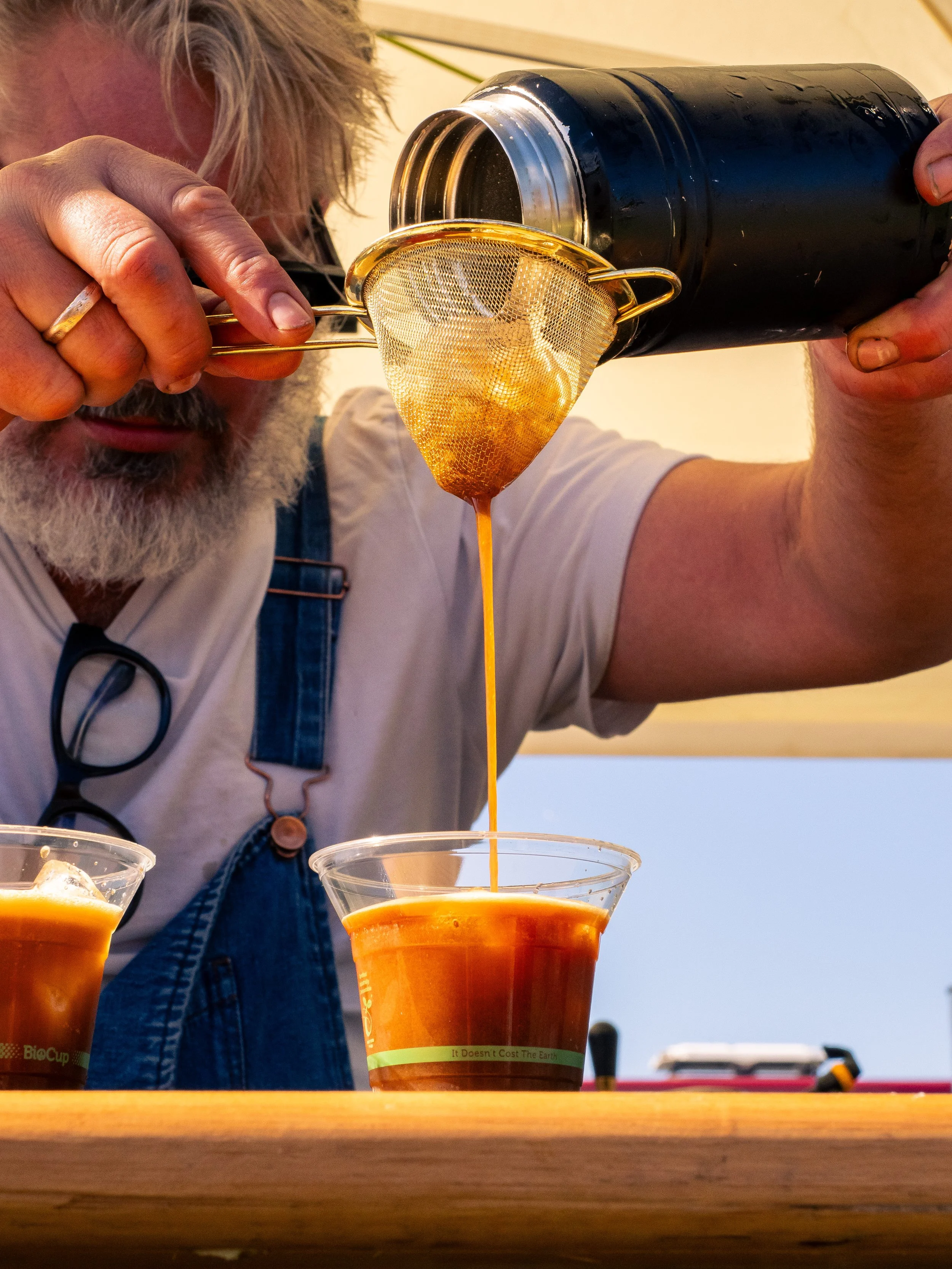 A man with gray hair and a beard is pouring orange liquid through a small mesh strainer into a clear cup. There are other cups with similar liquids on the table, and the man is wearing a white t-shirt and denim overalls.