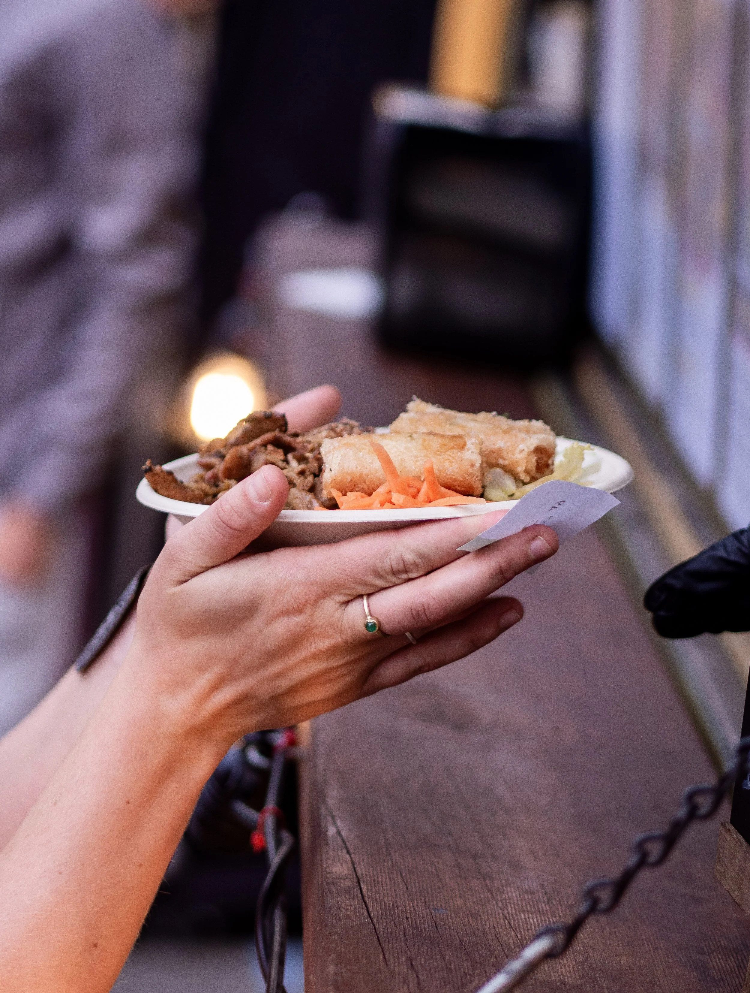 Close-up of two people holding a disposable plate of food with fried chicken, shredded carrots, shredded lettuce, and a biscuit, at an outdoor setting with chain links and a textured surface nearby.