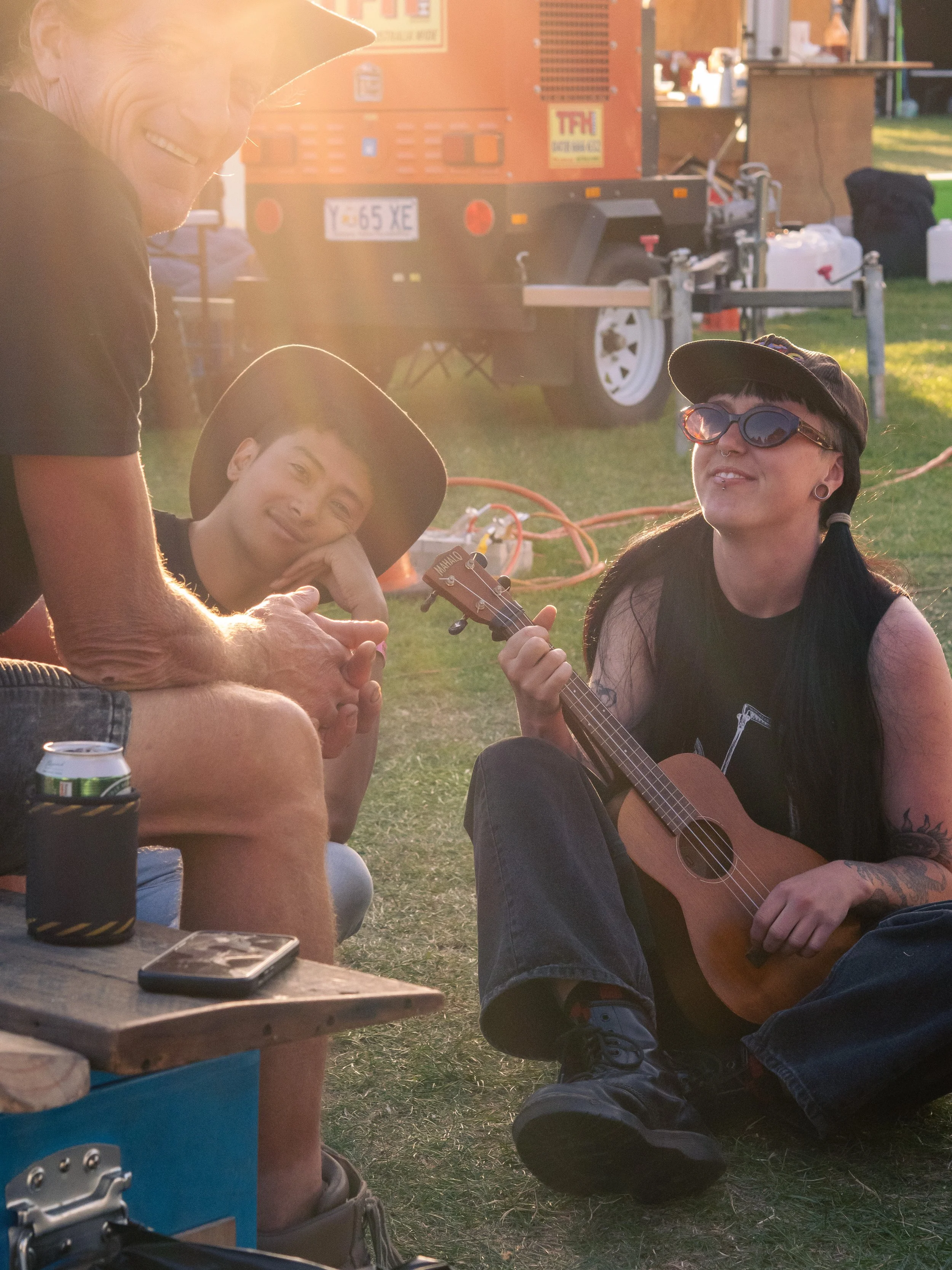 A woman with dark hair, tattoos, sunglasses, and a cap playing a small guitar outdoors, smiling and sitting on the grass during sunset. A young man with a hat, resting his chin on his hand, watches her. In the background, there's a trailers, containers, and a grassy area.