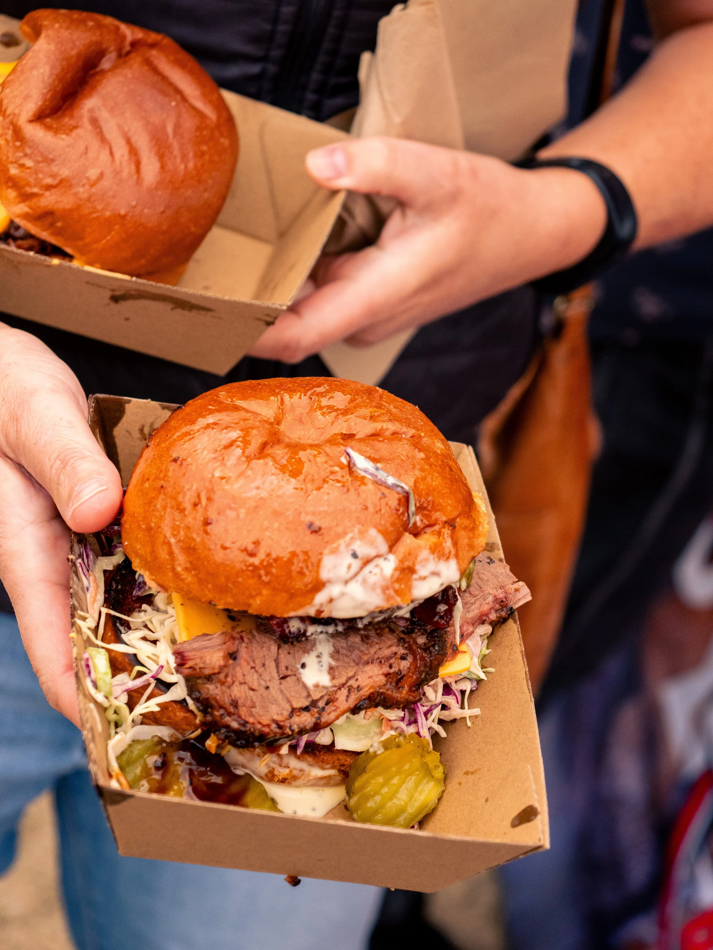 Close-up of a person holding a tray with a sandwich in a brown paper box, topped with a bun, shredded lettuce, and sliced meat, with pickles on the side.