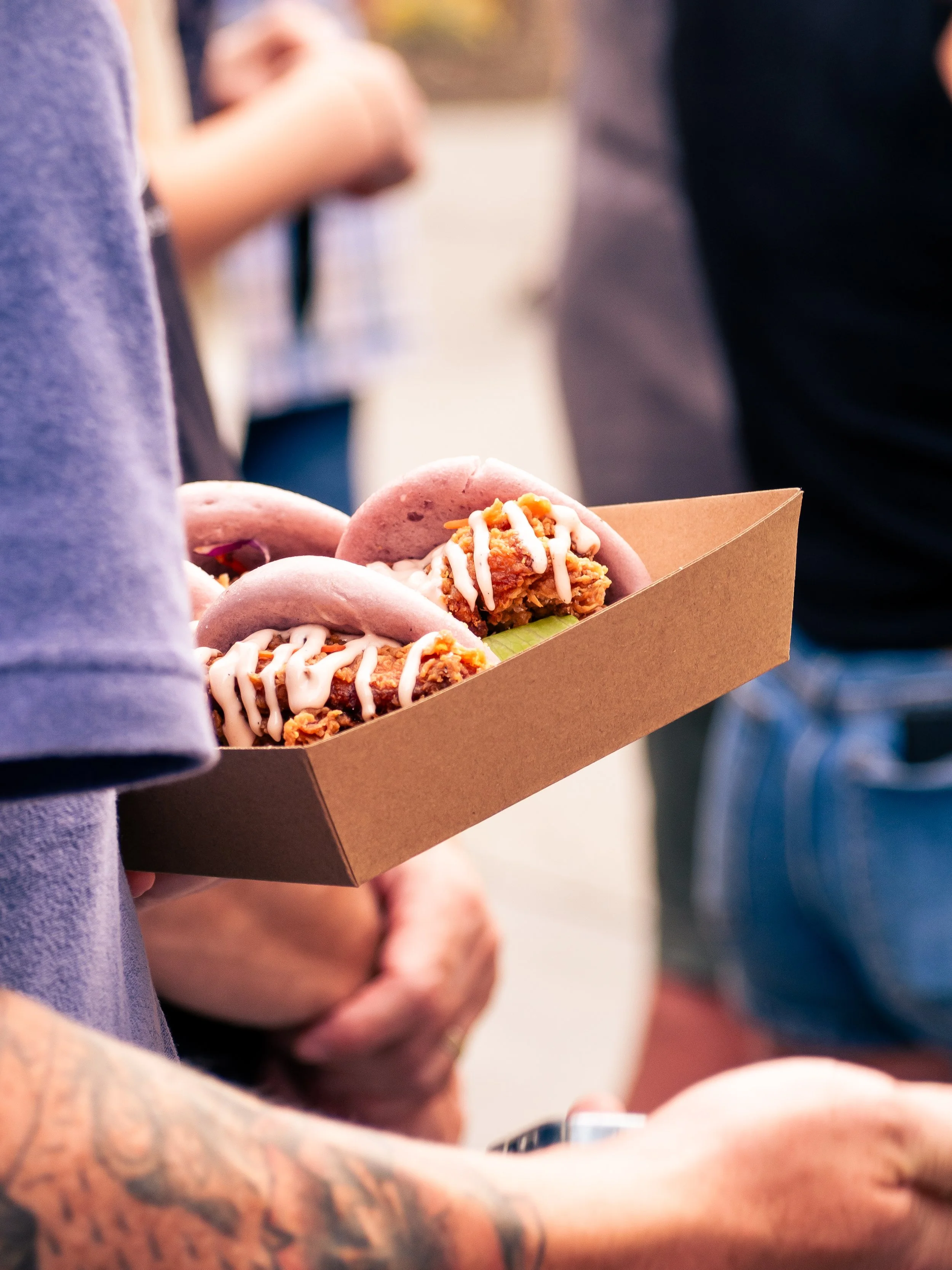 Person holding a cardboard tray of tacos topped with white sauce, with blurred people in the background.