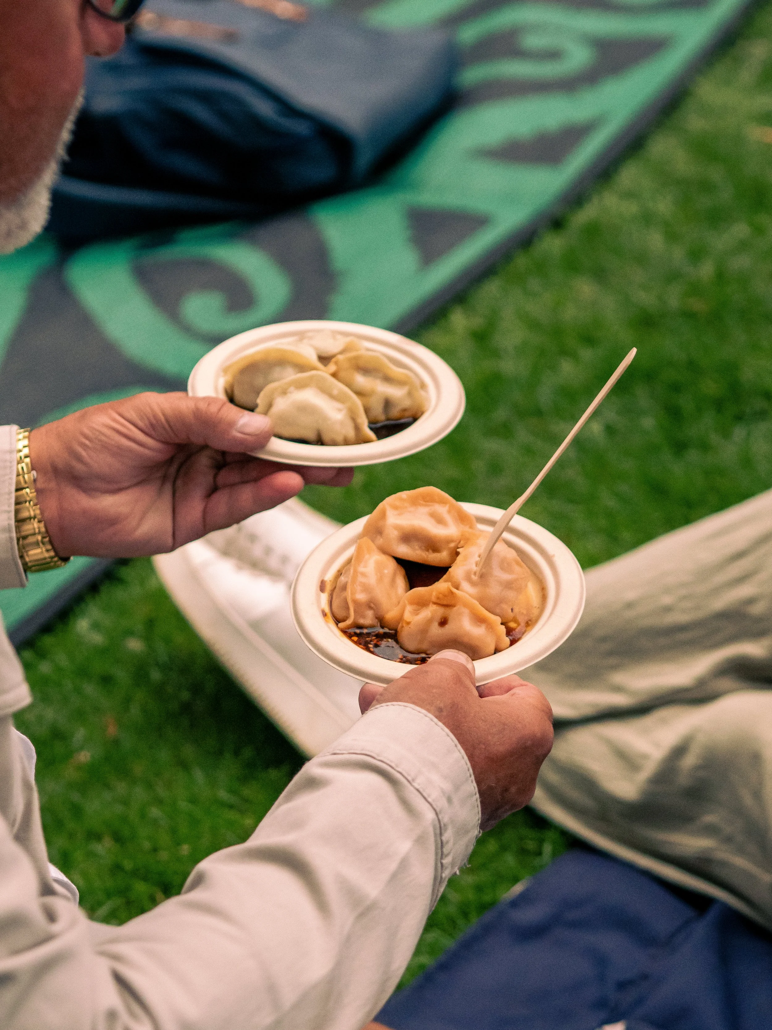 A person holding a plate of dumplings, with another plate of dumplings visible in the foreground, at an outdoor event.
