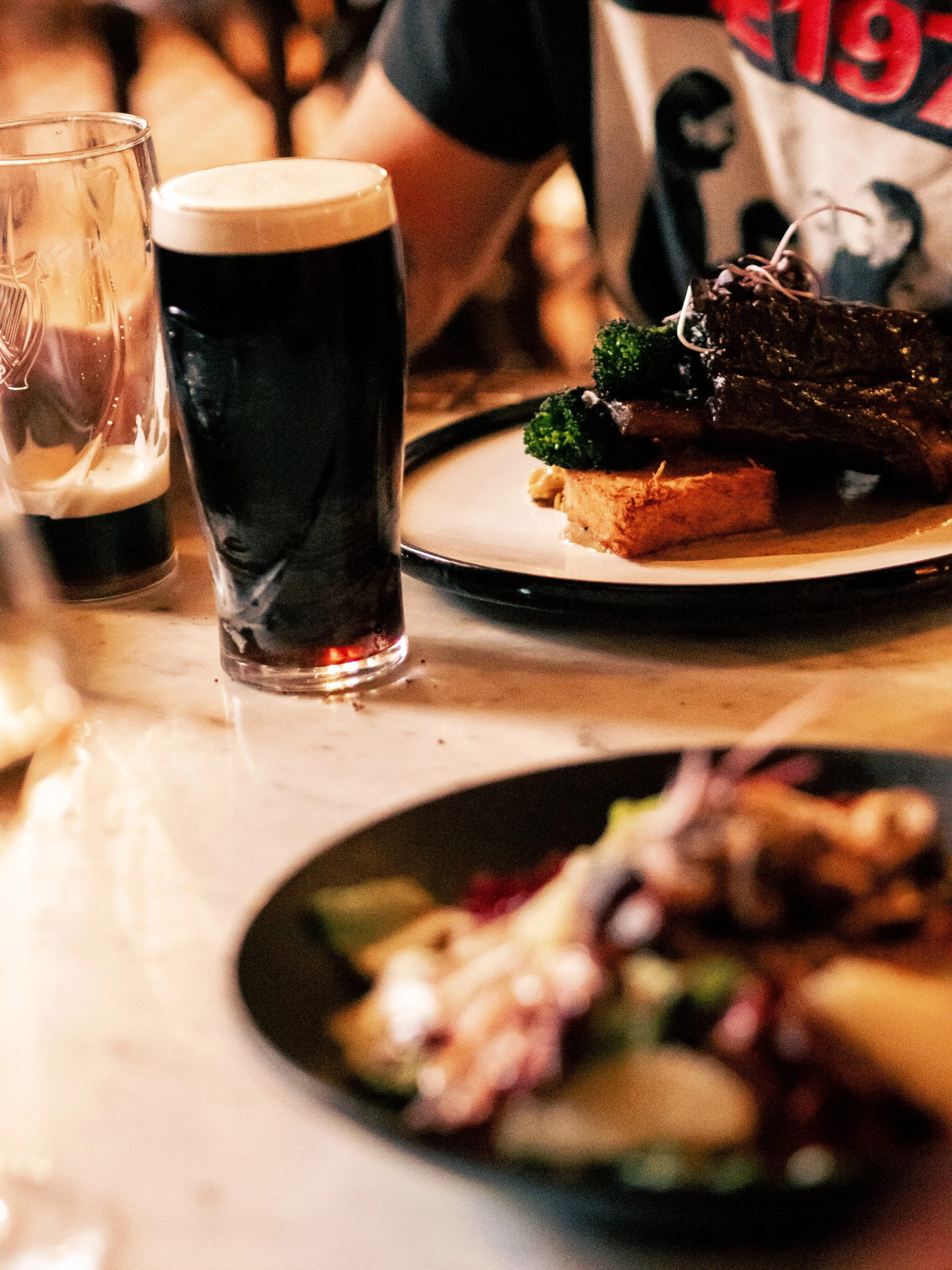 A plate of cooked steak with broccoli and mashed potatoes, two glasses of dark beer, and a person wearing a graphic T-shirt in the background.