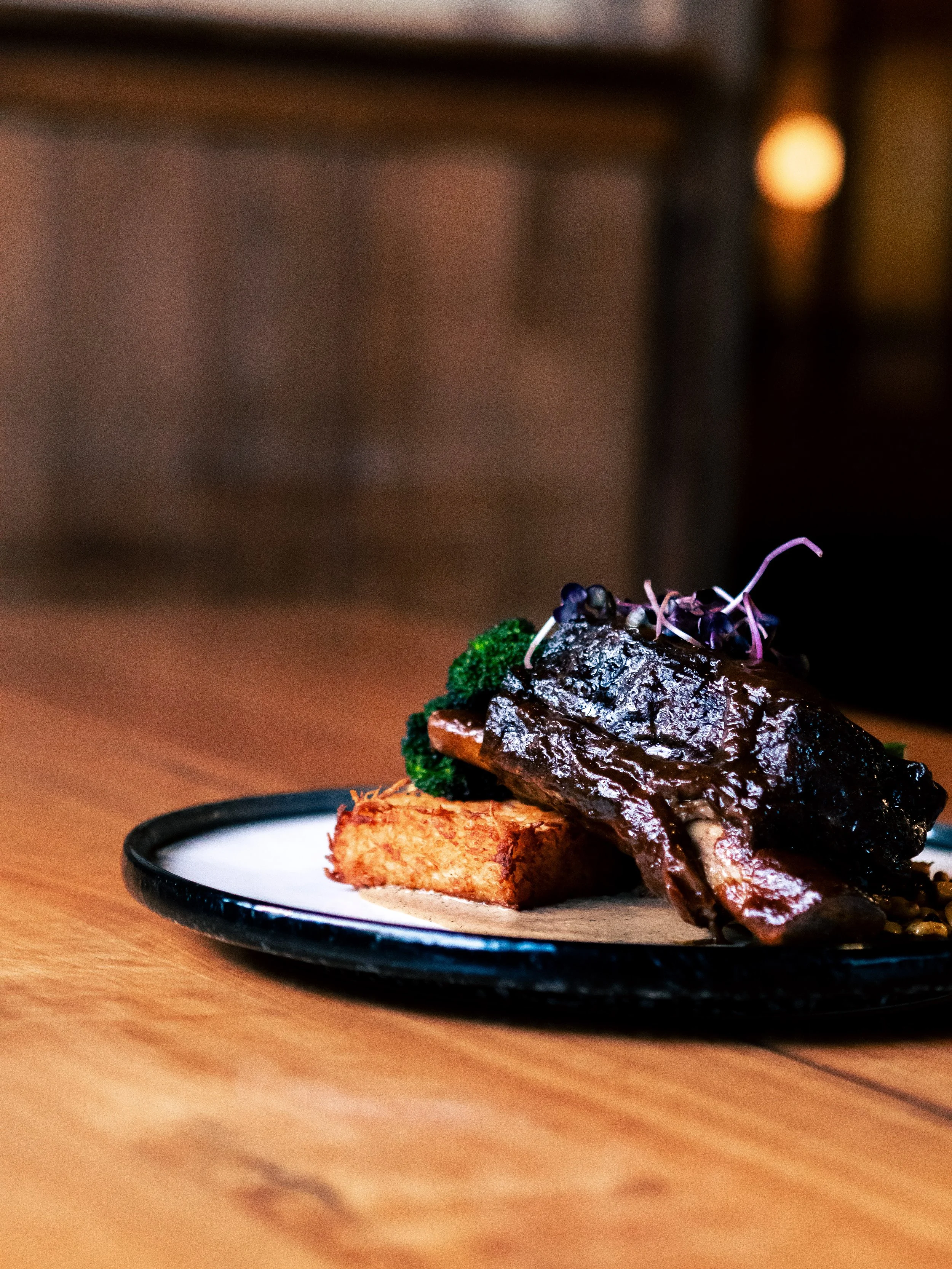 A plated serving of smoked beef rib with a layer of dark sauce, garnished with microgreens, on a wooden table.