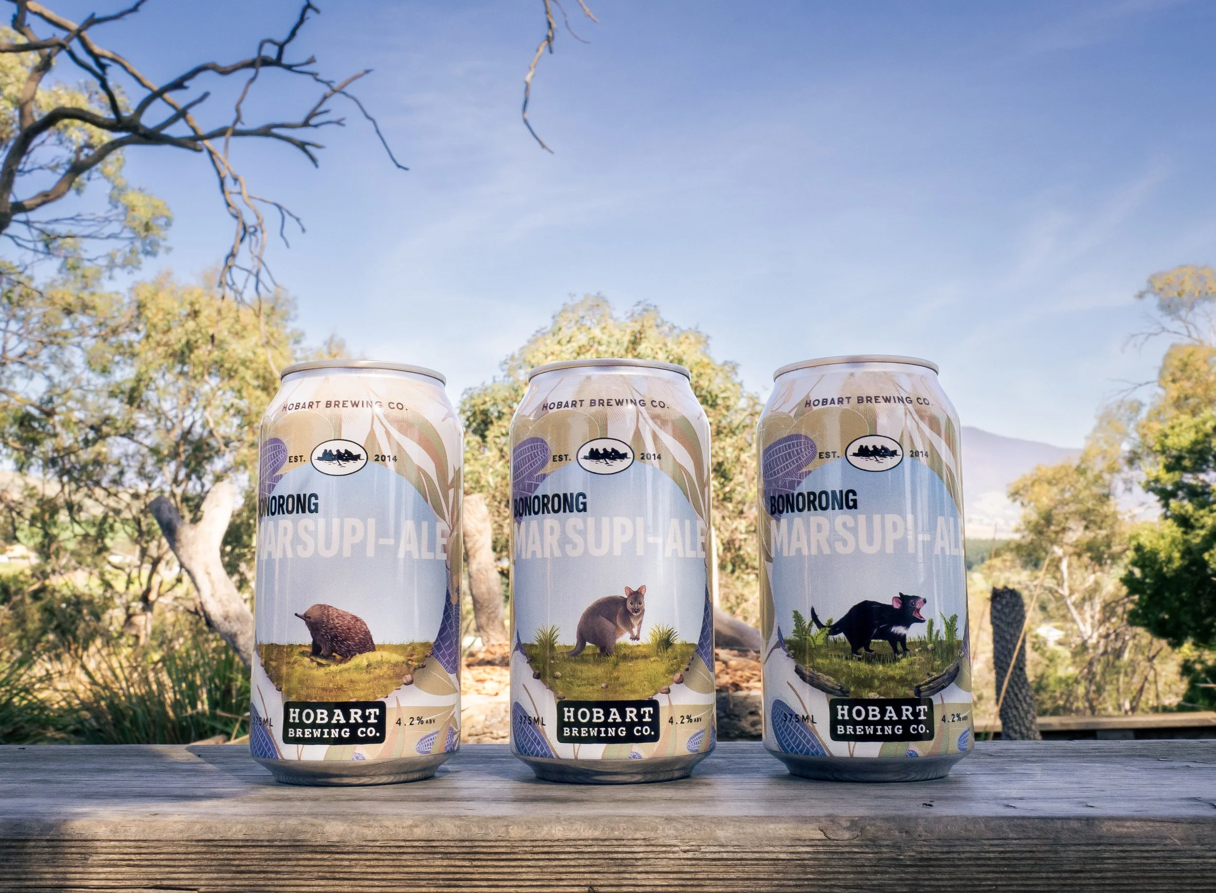Three cans of Hobart Brewing Co. Marsupi-Ale beer on a wooden surface outdoors with trees and blue sky in the background.