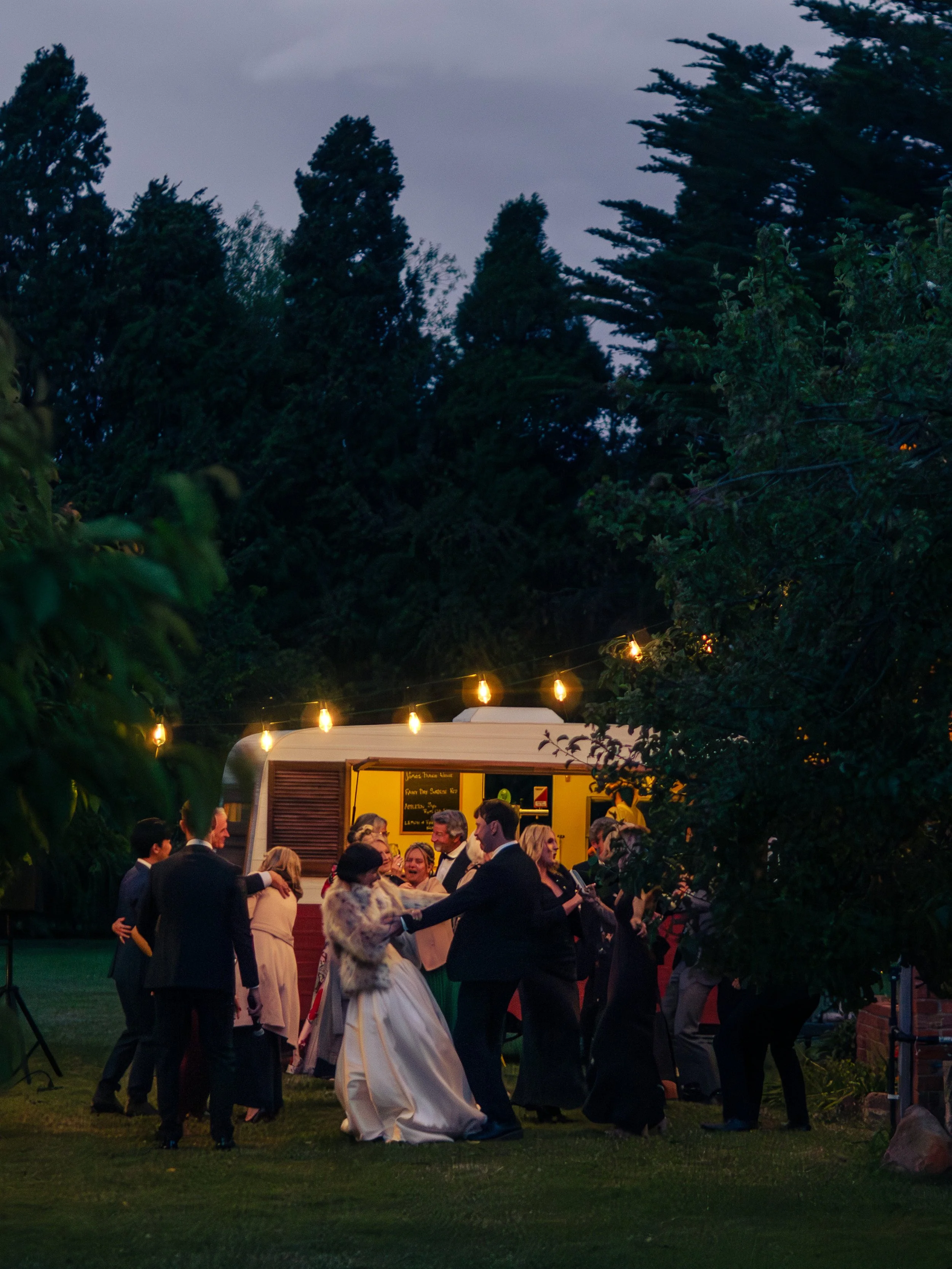 People dancing and socializing outdoors during evening event, with a food truck illuminated by string lights in the background, surrounded by trees.