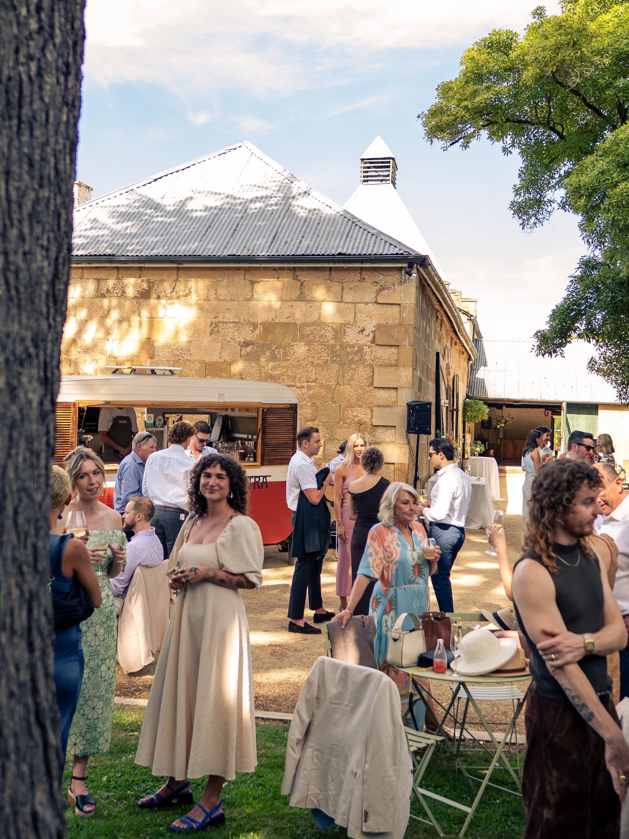 People gathered outdoors at a social event, with a rustic building featuring stone walls and a metal roof in the background, and surrounded by greenery and trees.