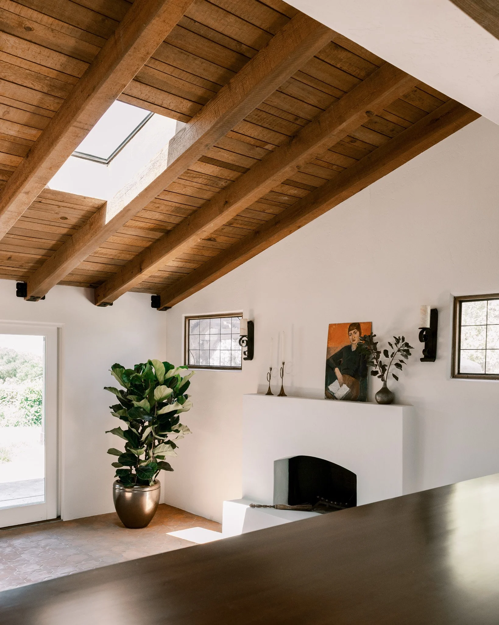 Interior of a bright living room with a white fireplace mantel, a potted plant with large green leaves, two small windows, a painting of a woman reading, and a fireplace with a fireplace tool in front.